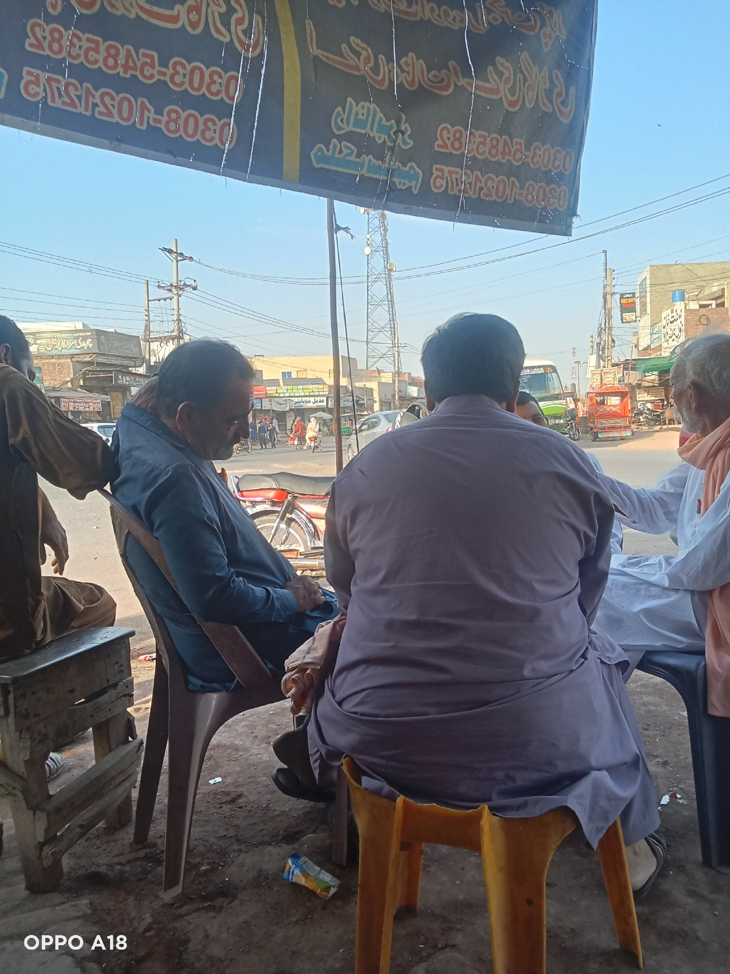 The image captures a street scene in what appears to be an urban setting, likely in an unknown city, based on the architectural style and the overall visual context. The composition focuses on a group of four men sitting in a shaded area, possibly a makeshift outdoor café or seating arrangement, suggested by the presence of stools and chairs. The man in the center is wearing a purple outfit and the man to the left of him is wearing a blue outfit, while the man to his right is wearing a white outfit. The fourth man is to the left and is wearing a brown outfit.
Several buildings are seen in the background, including some that may have commercial establishments, judging by signs or awnings visible. Traffic is also present in the background, with motorcycles, cars, and other vehicles moving along the road.
A sign hangs above the seated men, bearing Arabic or Urdu script along with telephone numbers. The scene appears to be a candid snapshot of daily life. The overall impression is that of a casual gathering in a busy and somewhat crowded urban environment.