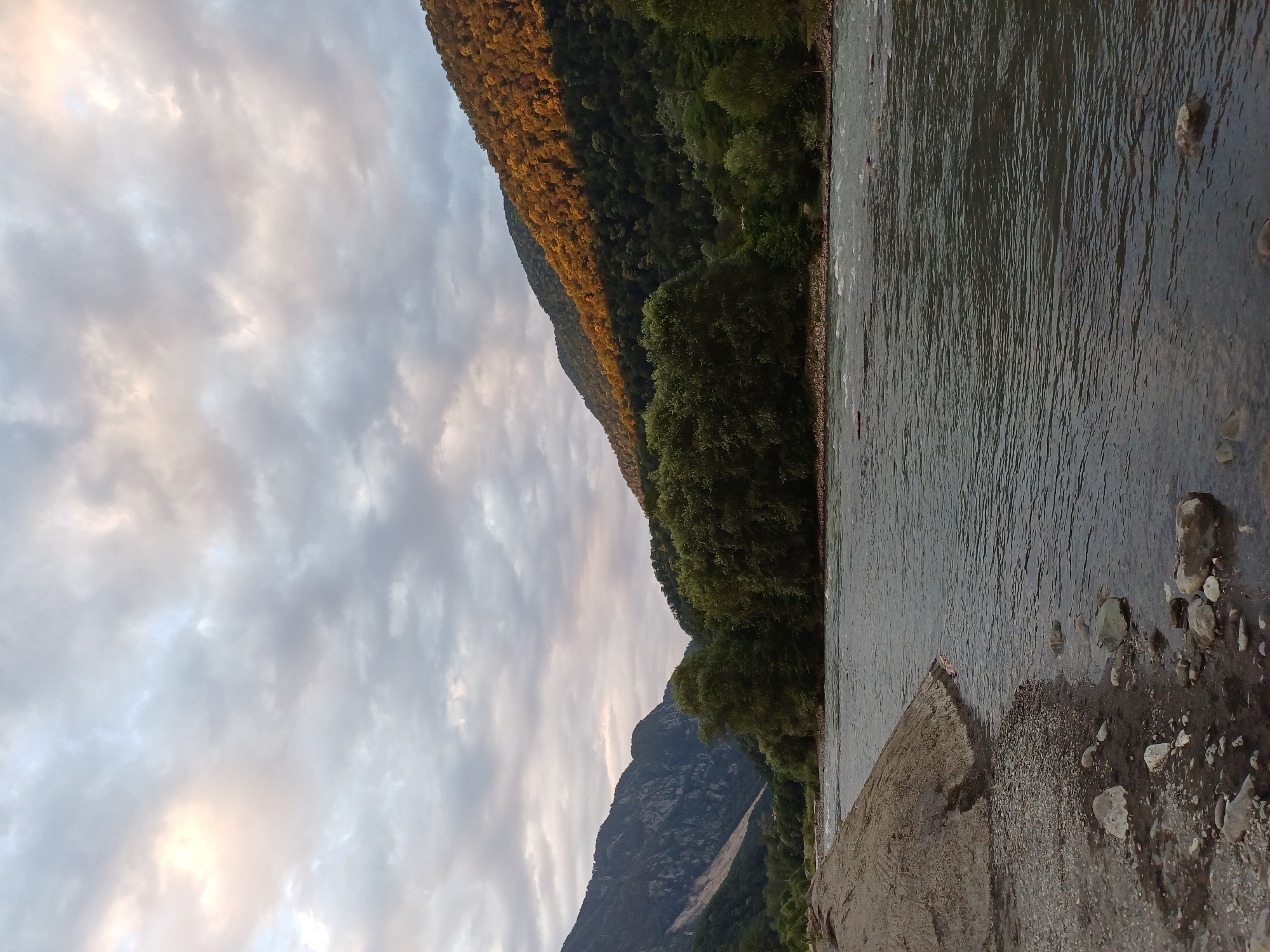The image depicts a natural landscape with a river flowing through a valley. Mountains covered in lush green trees flank the river. The sky is overcast with dramatic clouds, and the setting sun casts a warm glow on the upper slopes of the right mountain. The foreground features a rocky riverbank with pebbles and larger stones. The water appears calm, with some ripples indicating a gentle current. The scene is serene and untouched by human structures, suggesting a remote or natural area. Based on the visual cues of the mountainous terrain and vegetation, this scene is consistent with the natural beauty found in the vicinity of Adlerskiy, Russia, a region known for its mountainous landscapes and proximity to the Black Sea.