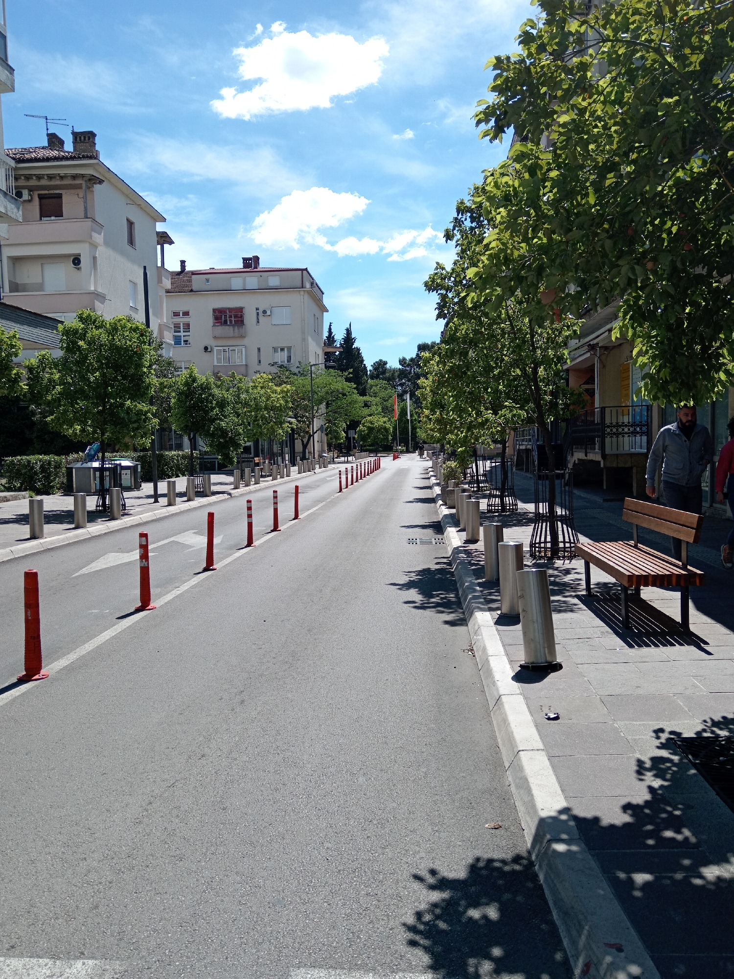 The image depicts a street in Podgorica, Montenegro on a sunny day. A straight, asphalt road stretches into the distance, marked by a white line and orange traffic bollards on the left. A white arrow is visible on the road pointing forward. On the left side of the road, there are silver bollards followed by trimmed trees. On the right side, a sidewalk is lined with trees in metal tree guards, silver bollards, and benches. A man in a gray jacket walks on the sidewalk. Buildings are visible on both sides of the street, along with a clear blue sky with some white clouds.