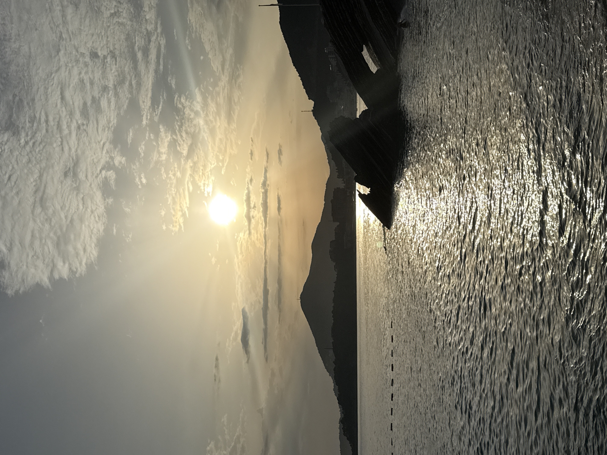 The image depicts a tranquil sunset scene at the coastline of Rafailovići, Montenegro. The sun, a bright orb, is positioned in the upper center of the sky, casting a warm, golden glow that reflects on the rippling surface of the calm sea. In the foreground, dark, jagged rock formations emerge from the water, creating a stark silhouette against the luminous sea and sky. The middle ground is occupied by the calm sea, with a series of small, dotted buoys forming a dashed line across the water. Beyond the sea, a silhouetted landscape of rolling hills and mountains stretches across the horizon. Some structures, possibly buildings, are faintly visible on the slopes of the hills. The sky is a gradient of soft grays and yellows, with scattered clouds tinged by the setting sun. The overall atmosphere is serene and picturesque.