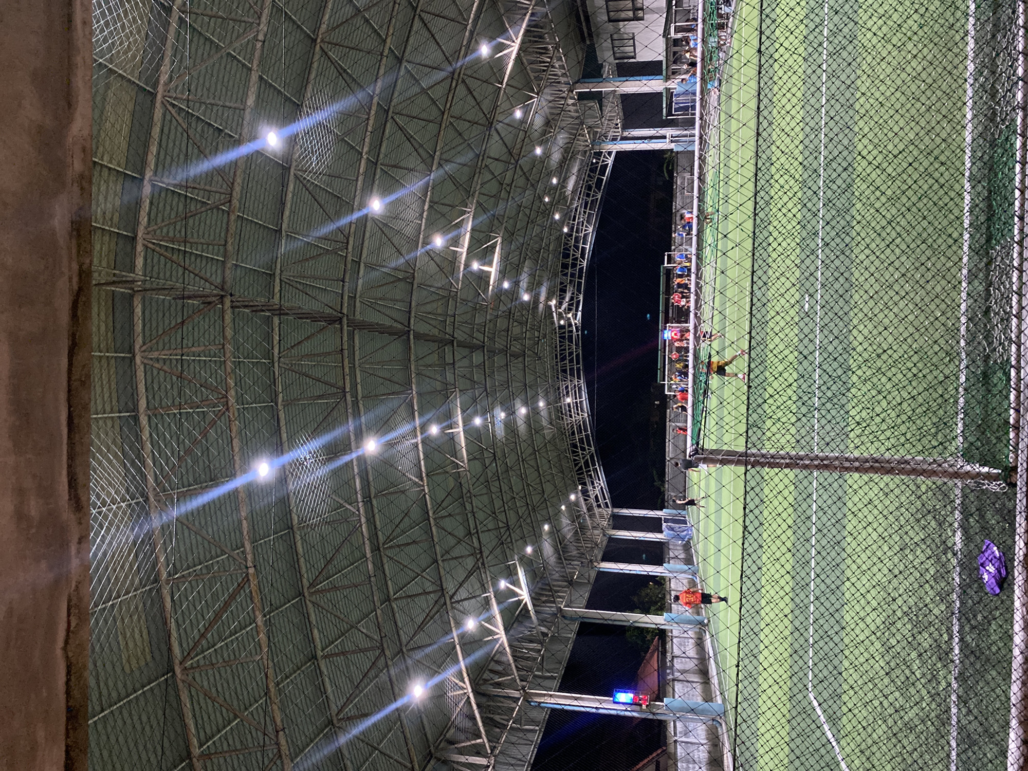 This image shows an indoor soccer field in Baan Chaweng Noi, Thailand. The field is enclosed by a high net. People are seen on the sidelines and playing on the field. The lighting consists of bright, spaced-out ceiling lights. The roof structure is a metal framework. A multi-story building is visible in the background.