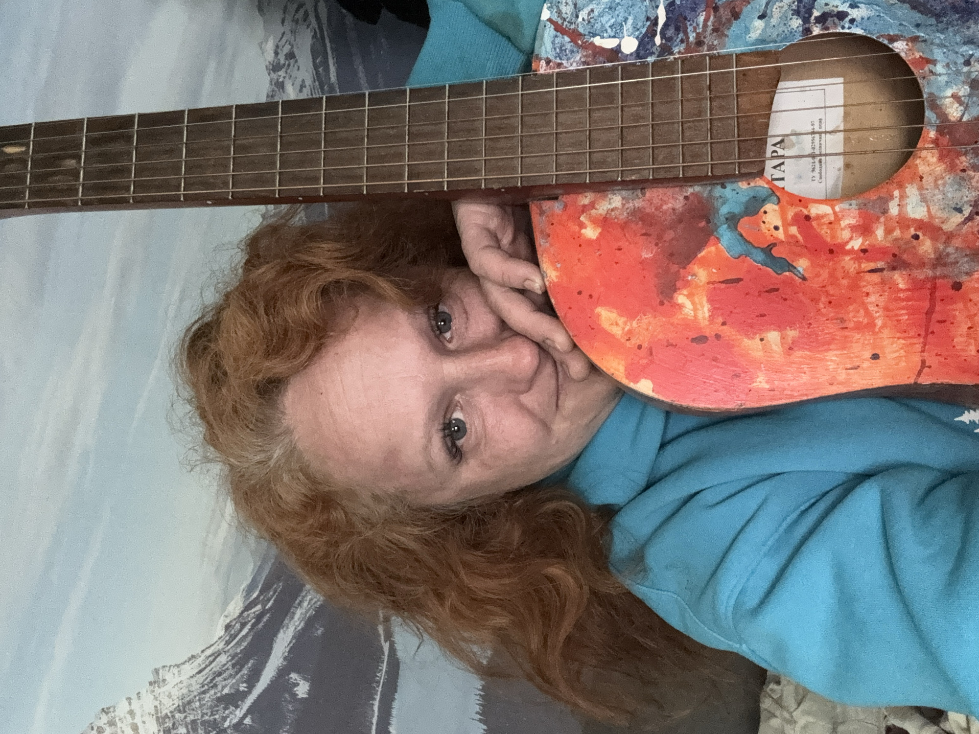 A woman with red curly hair is lying on her side, wearing a blue hoodie, with a painted acoustic guitar resting next to her face. The guitar has a red and blue paint design. The guitar's fretboard is positioned above her head. Behind her is a surface with a gray and white mountain range design. The interior label on the guitar reads "TAPA". The image was taken in Chaikovsky, Russia.