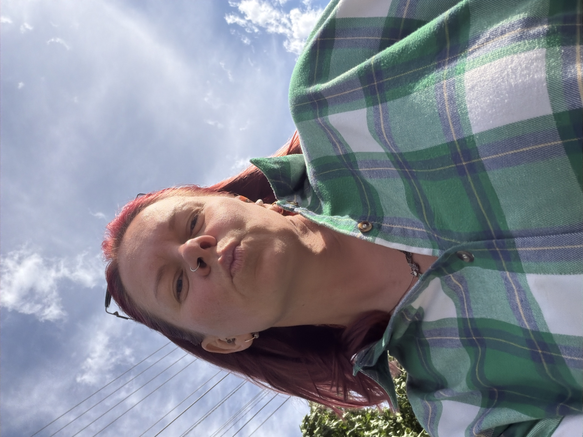 A woman is depicted in a selfie, tilted, wearing a green and white plaid shirt. She has red hair, a nose ring, and earrings. The background shows a partly cloudy sky and power lines. Some green foliage is visible. The location in the city of Chaikovsky, Russia, cannot be determined from this image.
