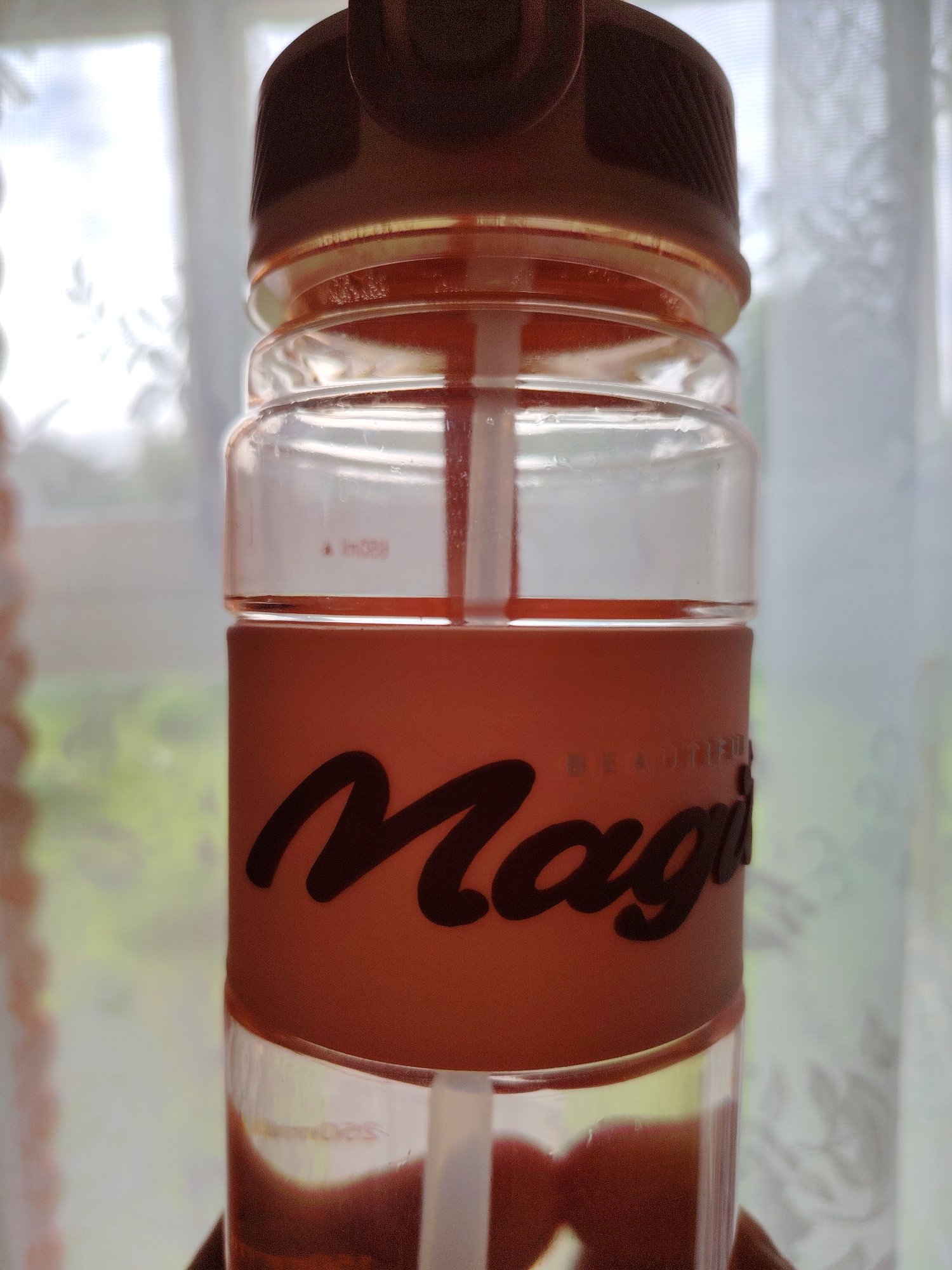 The image shows a close-up of a clear plastic water bottle with a dark brown lid and a light orange band around the middle. The band has black cursive lettering that reads "Magic" and a smaller phrase "BEAUTIFUL" above it. A white straw is visible inside the bottle, extending from the lid towards the bottom. The bottle contains a reddish-brown liquid, possibly a beverage. The background is out of focus, showing a window with white curtains and blurred greenery outside, suggesting an indoor setting with natural light. The image does not contain any elements that would allow for identification of the location within the city of Soivolovskaya, Russia.