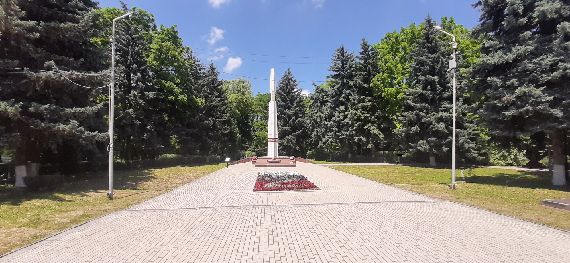 The image captures a war memorial located in the city of Yessentukskaya, Russia. The memorial features a tall, white obelisk with red accents, situated on a raised platform. In front of the obelisk is a rectangular flower bed filled with red and white flowers. The memorial is surrounded by a paved walkway and a grassy area with trees, primarily coniferous and deciduous varieties, providing a lush green backdrop. Two tall lamp posts stand on either side of the walkway, flanking the approach to the memorial. The sky above is blue with scattered white clouds, indicating a clear day. No people are visible in the scene.