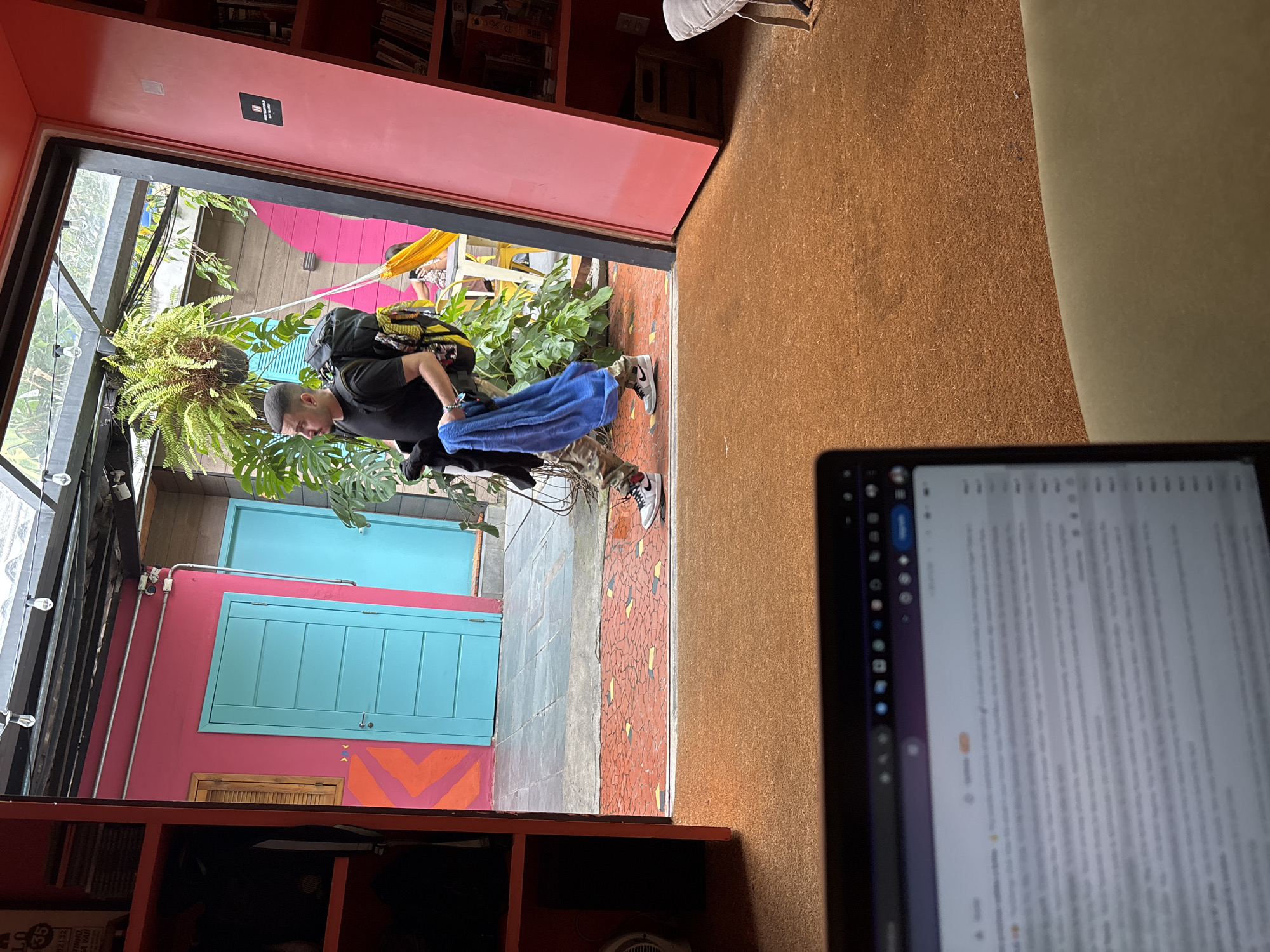 A view from inside a room in the city of São Paulo, Brazil, through a doorway to an outdoor space with colorful architecture. A person wearing a black shirt and carrying a backpack and blue towel is walking out the doorway. The architecture outside the doorway includes pink walls, blue doors, a glass ceiling, and a vibrant display of plants. Inside the room, there is a red bookshelf on the left and a laptop screen in the foreground on the right, displaying text. The room has a brown wall.