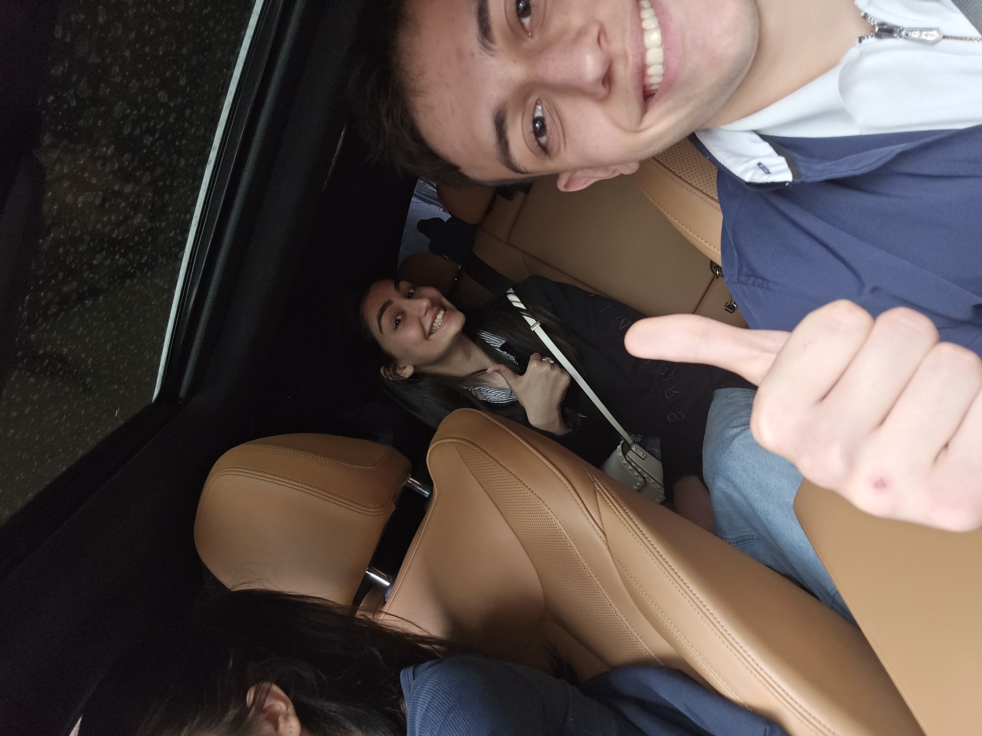 Inside a car, a man in a blue jacket and light blue jeans points at the camera with his right index finger while smiling. A woman sits in the back seat, also smiling. The car has tan leather seats and a sunroof. The location appears to be Yerevan, Armenia.