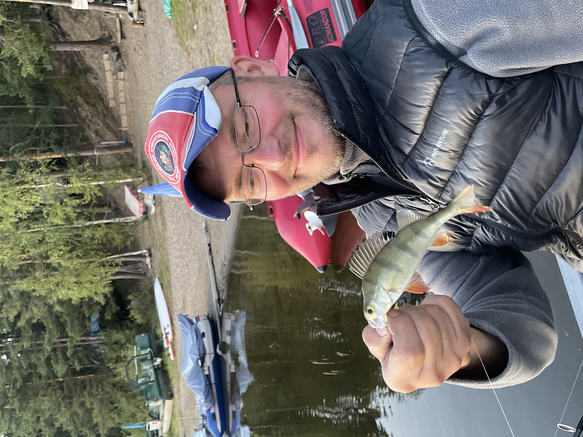 In Berozovo, Russia, a man wearing a puffer vest, a grey fleece jacket, and a red, white, and blue baseball cap smiles at the camera while holding a small, freshly caught fish in his left hand. He is positioned in the foreground, with a body of water behind him, reflecting the surrounding trees. Several inflatable boats are visible near the shore, along with some sandy beach area and dense forestation in the background. The man's fishing line extends from the fish to his right.