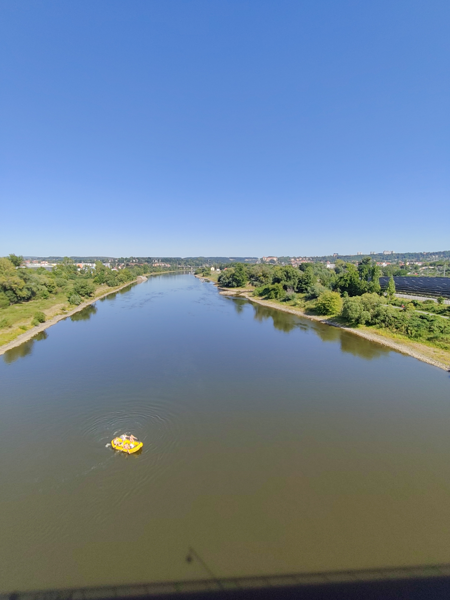 An aerial, high-angle, vertical shot captures a wide, calm river flowing through the city of Pirna, Germany, under a clear blue sky. A small, bright yellow inflatable raft with at least three people is positioned in the left third of the river, its occupants appearing relaxed as they float. The riverbanks are lined with lush green trees and vegetation, with a town visible in the distance on the right bank, characterized by buildings and what appears to be a solar panel farm. The overall scene conveys a peaceful summer day with recreational activity on the water.