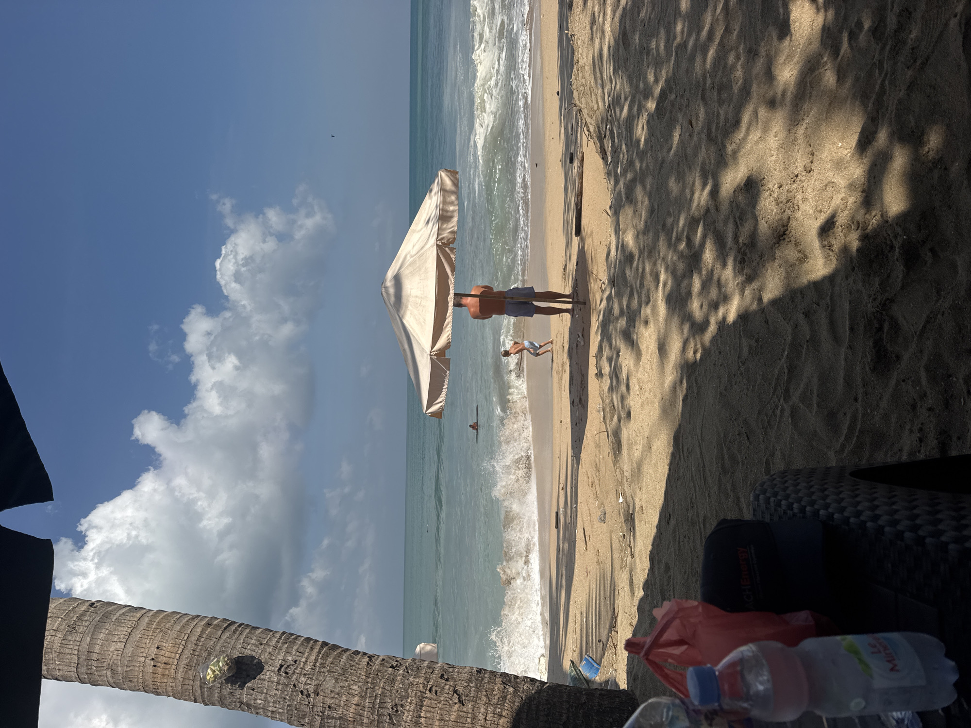 In Kuta, Indonesia, on a sunny day at the beach, a person stands under a large umbrella, facing the ocean. To the left, a coconut tree trunk is visible, with a plastic bag containing some items attached to it. In the foreground, a table with a water bottle and a red bag rests on the sand. A child is playing in the shallow water near the shore, while waves crash on the beach. The sky is blue with scattered clouds, and the water is a turquoise color. There are shadows cast on the sand by the umbrella and palm tree, indicating the time of day. The scene is one of a typical beach day, with people enjoying the sun and sea.