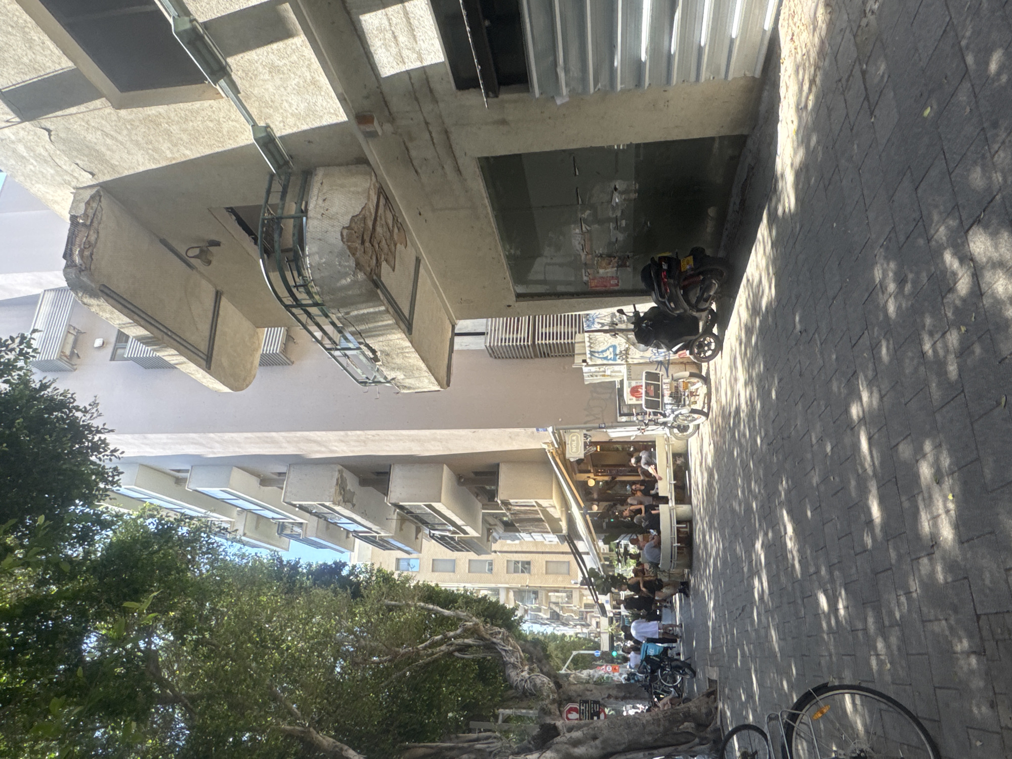 This image depicts a street scene in Tel Aviv, Israel. Along the right side of the frame is a paved sidewalk covered in dappled sunlight. A bicycle is parked, its wheels visible at the bottom of the frame. Adjacent to the sidewalk, storefronts are visible, with people seated at tables under awnings, suggesting outdoor dining at a cafe or restaurant. Further down the street, more people are walking and biking. A row of buildings lines the left side of the frame, with trees providing greenery. The perspective is slightly tilted.