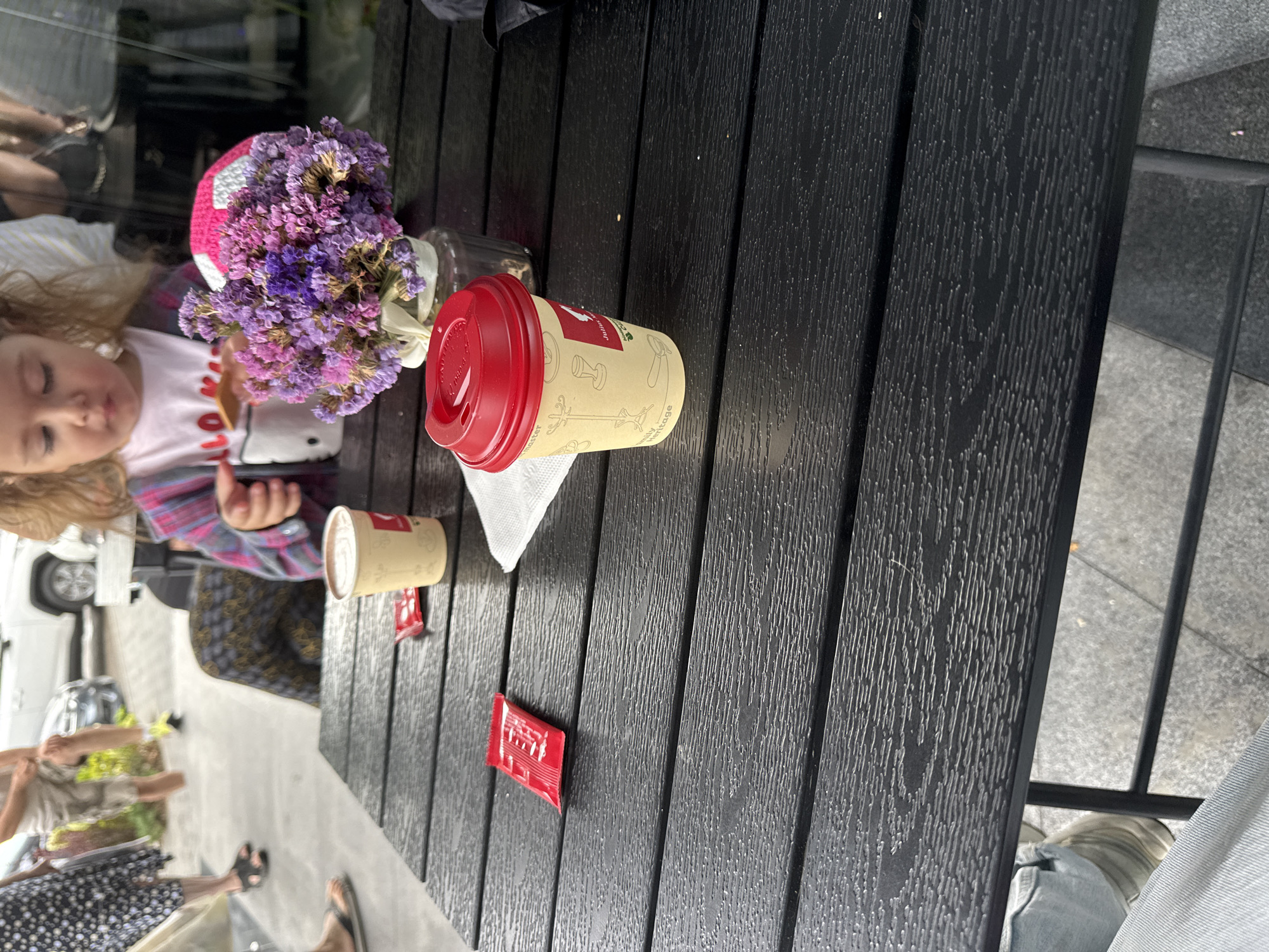 On a dark, textured wooden table, two disposable coffee cups with red lids are present. One cup, on the right, is a light cream color with a red and white logo and coffee-themed illustrations. It sits next to a white napkin. A small bouquet of dried purple flowers in a clear glass vase is positioned behind this cup. To the left of this cup, a second, slightly taller, cream-colored cup is visible, also with a red lid. A small red packet, possibly sugar or creamer, lies on the table between the two cups. In the background, a young girl with blonde curly hair wearing a pink and plaid shirt is partially visible, reaching for something on the table. Another person, a woman in a black and white patterned dress, stands in the background to the left, as does a man in shorts and a light-colored shirt. The scene appears to be outdoors, possibly a cafe or restaurant patio in Chișinău, Moldova, given the context provided.