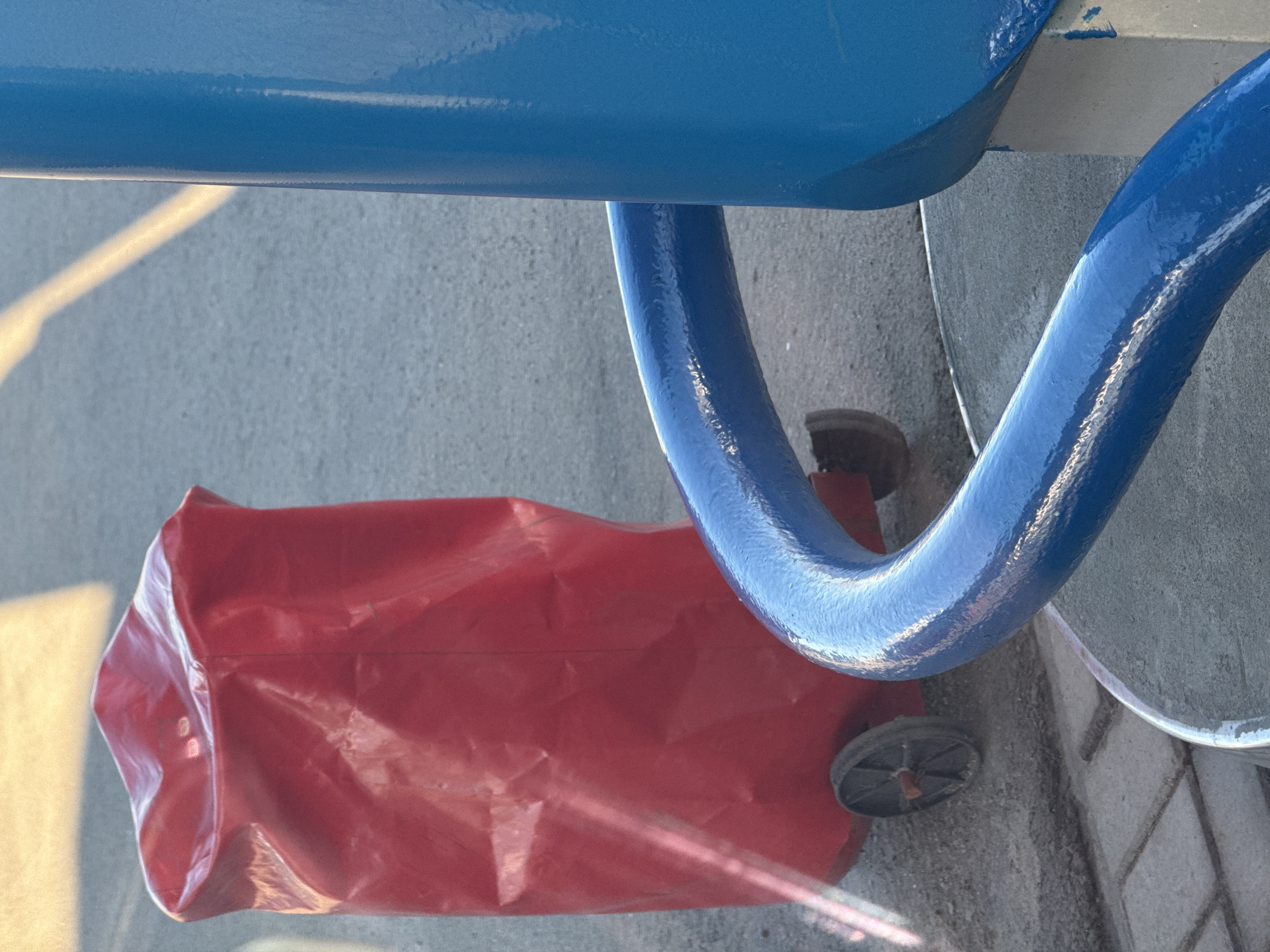 The image depicts a red shopping bag with wheels resting on a gray concrete surface. Overhanging the bag are sections of a blue, possibly metal, structure, which could be part of a fixture or street furniture. The light suggests the photograph was taken during the day. There are no people visible. The location is not discernible from the image and therefore cannot be verified as Ново-Никольское, Russia.