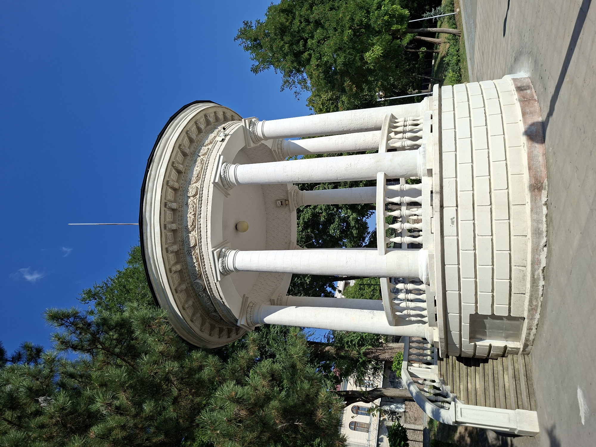 A white, classical rotunda with a balustrade and Ionic columns stands on a paved area under a clear blue sky. Tall green trees, including a pine tree on the left, surround the rotunda. A set of stone stairs with a white balustrade leads up to the rotunda on the left. In the background, a building with arched windows is visible. The ground is paved with light gray concrete, and a shadow of a pole and its light fixture is cast on it. The rotunda is adorned with decorative carvings along its upper edge. Some graffiti is present on the columns of the rotunda.