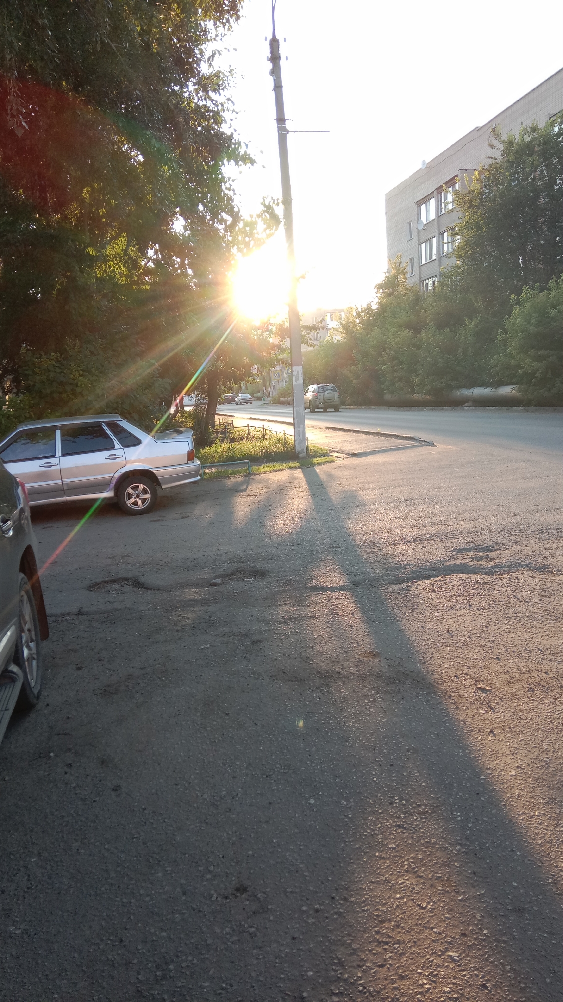 In Petropavl, Kazakhstan, a street scene unfolds bathed in the warm glow of the setting sun, casting long shadows across the asphalt. A silver sedan is parked on the left side of the image, its metallic surface reflecting the golden light. A street lamp stands tall, its pole casting a distinct shadow that stretches diagonally across the road. Beyond the lamp, a multi-story building with rows of windows is visible, partially obscured by lush green trees. Further down the street, another vehicle, an SUV, is driving away from the camera. The road itself shows signs of wear, with small potholes visible. The sky is bright and clear, indicating a sunny day.