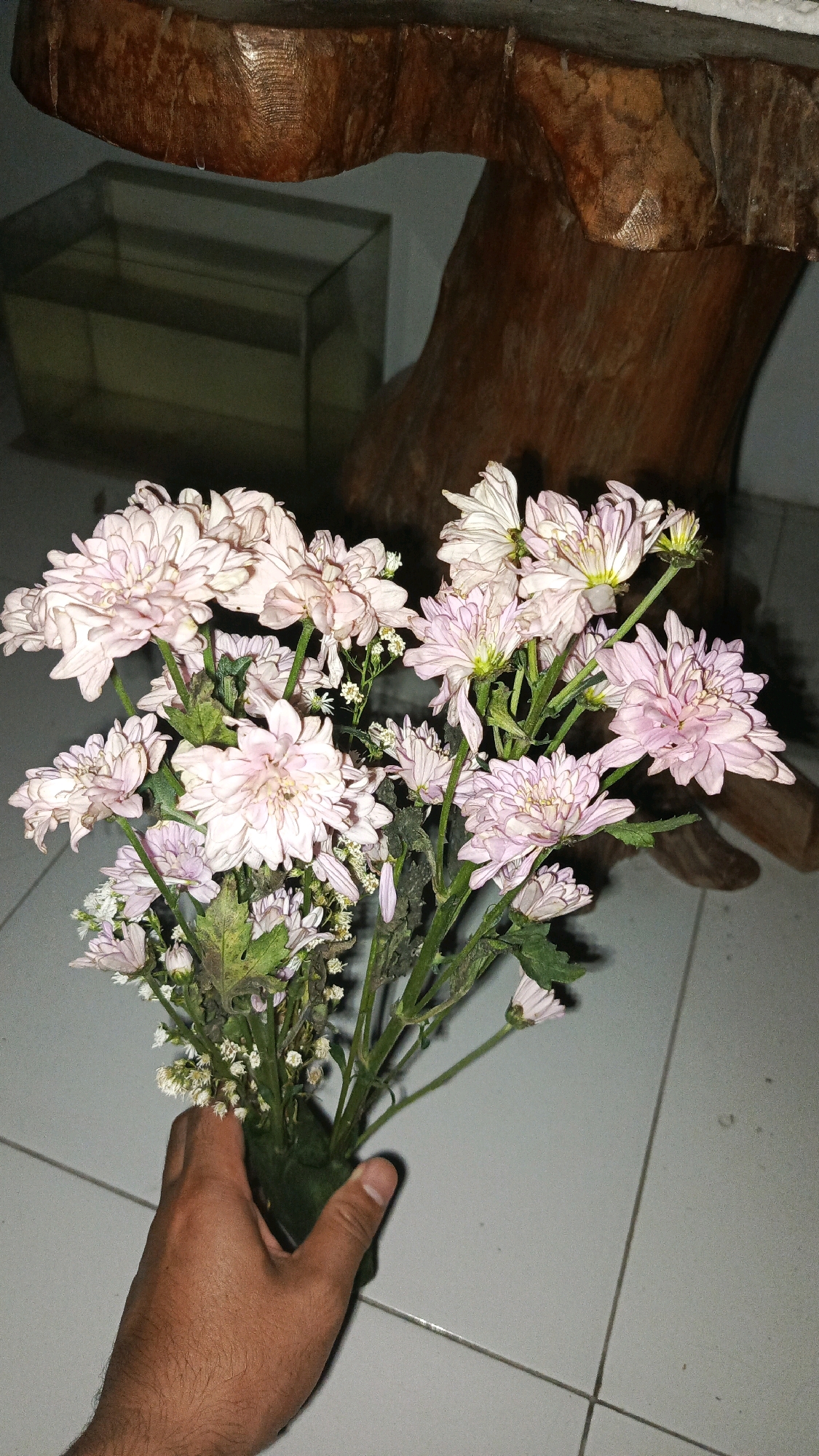A person's hand holds a bouquet of light pink and white flowers, some with yellow centers, against a tiled floor and a blurred background featuring a wooden structure and a glass aquarium. The lighting suggests an indoor, possibly nighttime, setting in Babadan, Indonesia. The flowers are varied in size and bloom, with some appearing slightly wilted. The wooden structure appears to be part of a table or decorative piece, with a visible grain. The aquarium contains a yellowish liquid and is positioned behind the flowers. The floor is composed of square tiles with grout lines.