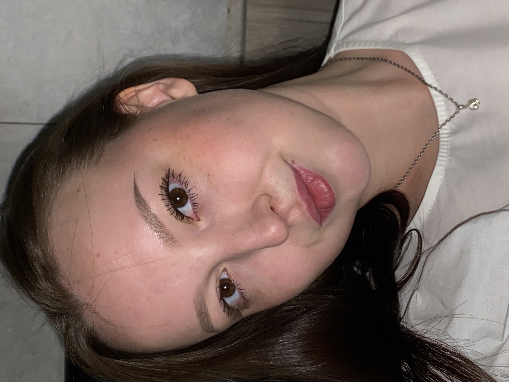 A close-up shot of a young woman with brown hair, wearing a white shirt and a silver necklace with a small diamond pendant. She is facing upwards with her head tilted slightly to the left. Her skin appears fair, and she has brown eyes, light eyebrows, and pink lips. She appears to be wearing mascara on her eyelashes. The background is indistinct, but appears to include a wall. There is no explicit evidence that indicates the location is Chaikovsky, Russia.