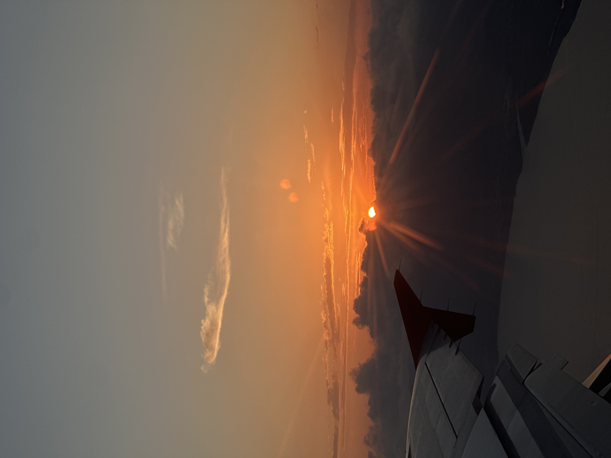 An aerial photograph captures a sunset viewed from an aircraft. The upper sky displays a gradient from deep grey-blue at the zenith to a vivid orange and yellow near the horizon. The sun, partially obscured by clouds, is visible as a bright orange orb, emitting prominent crepuscular rays that fan outwards. A band of dark, voluminous clouds lies below the sun, largely silhouetted, while wispy, elongated clouds are present higher in the frame. The left and lower portions of the image feature the dark, metallic wing of an airplane, seen from an interior perspective, with its flaps and a red wingtip discernible. Below the wing and the lower cloud layer, a dark body of water is visible, bordered by a faint, dark landmass. No people are directly visible. The scene is consistent with a flight near the city of Dalaman, Turkey.