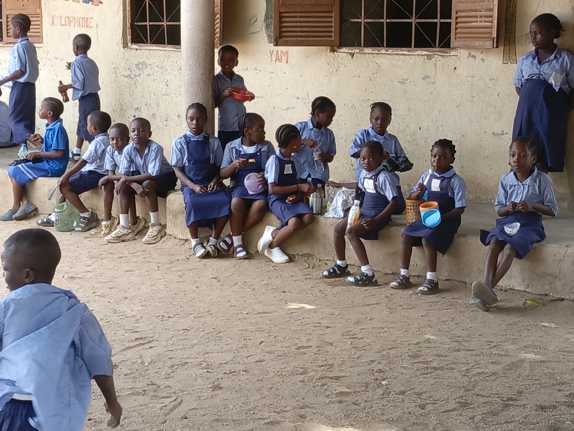 The image depicts a group of children, likely students, gathered outdoors at what appears to be a school in Akwanga, Nigeria. The setting features a dusty, unpaved ground surface and a light-colored building in the background with visible windows, some fitted with wooden shutters, and a large supporting pillar.
Most of the children are seated on a low concrete ledge or platform against the building. They are dressed in light blue short-sleeved shirts and dark blue skirts or shorts, indicating school uniforms. A central group of girls is seen holding various items such as small plastic bowls (in red, purple, and blue), water bottles, and a metal thermos, suggesting they are having a snack or meal. Some children look directly towards the camera, while others are engaged in eating or observing their surroundings.
On the left, several boys are also seated on the ledge. In the foreground, a boy walks away with his back to the camera. In the background, other children are standing or moving. The scene conveys a moment of recess or lunch break.
The weather appears sunny with bright natural light. Visible text painted on the building wall includes "X LOPHINE" (likely part of "Xylophone") and "YAM". The overall atmosphere is relaxed, with children exhibiting calm expressions.