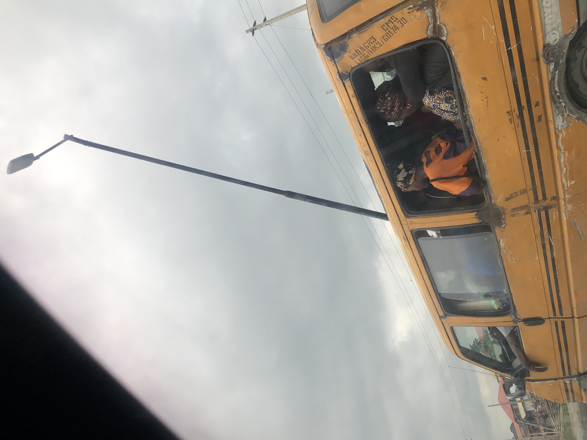 A yellow minibus, likely a commercial transport vehicle, is shown from a side angle under a cloudy sky. The van's body is weathered and shows signs of rust and repair. Several passengers are visible through the open windows, with one person's arm resting on the driver's side window frame. Text is visible on the side of the van, including "BADAGRY," "CMS," "LASG/HKG/60174.30". A tall street lamp pole extends diagonally across the frame, reaching towards the top right corner of the image. In the background, there are some buildings and a utility pole with power lines. The scene appears to be in an urban or semi-urban area, possibly along a roadside in Satellite Town, Nigeria, given the text on the vehicle.