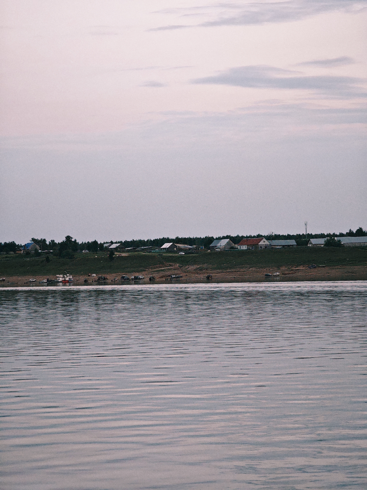 This is a horizontal, wide-angle shot of a village on the bank of a body of water. In the foreground, the water is calm with gentle ripples reflecting the soft, diffused light of a cloudy sky. In the middle ground, a village is nestled on a grassy incline dotted with trees. Several houses with various colored roofs are visible, and a line of vehicles, including cars and what appear to be utility vehicles, are parked along the water's edge. A small boat or barge is also present in the water. Behind the village, a dense line of dark green trees forms a backdrop. The sky above is a pale, hazy pink and blue, suggesting either early morning or late evening. The overall mood is serene and peaceful. There is no visible text in the image.