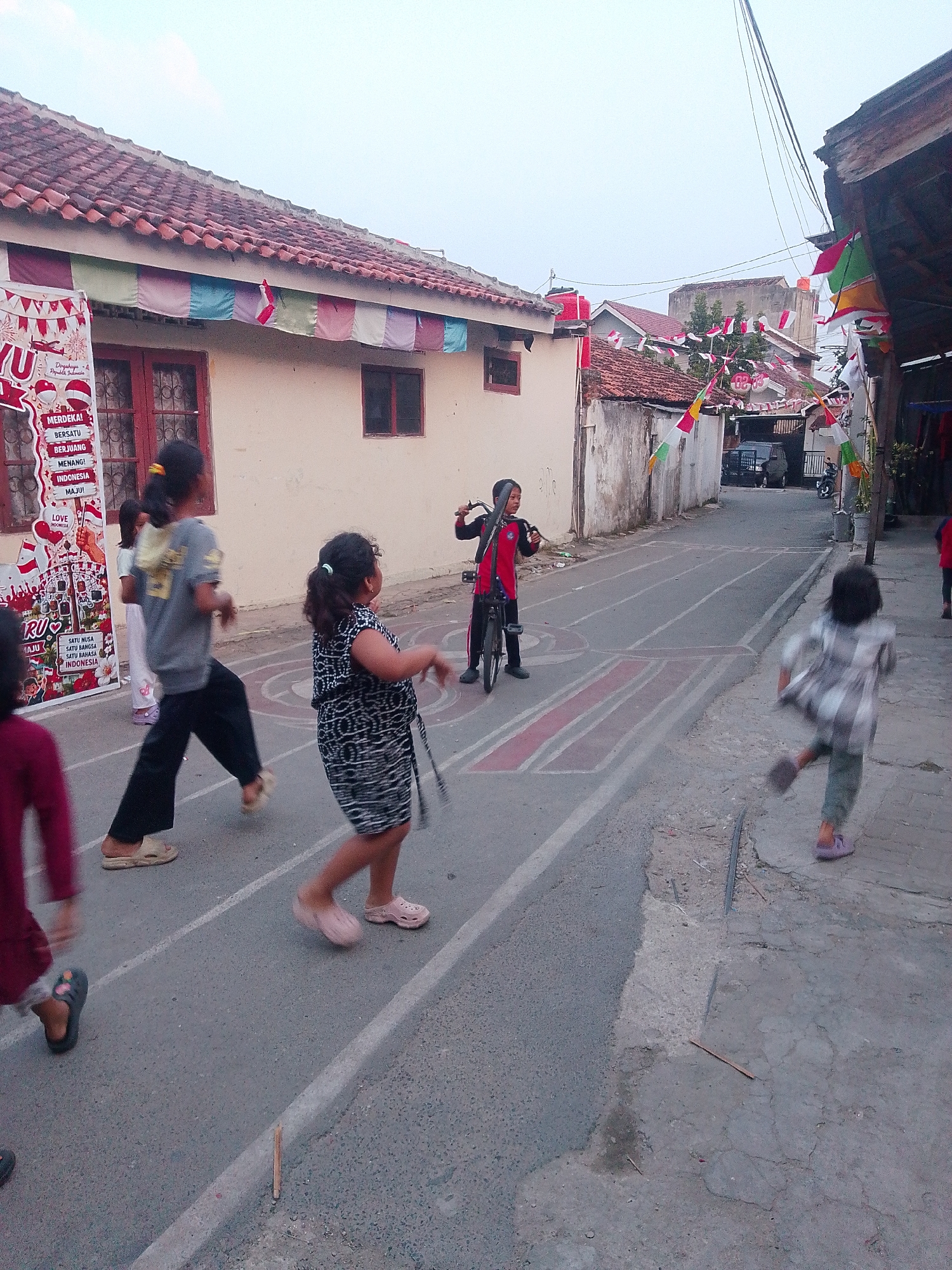 In the city of Bandar Lampung, Indonesia, a group of children is engaged in various activities on a paved street lined with houses decorated with Indonesian flags. One boy in a red and black outfit is on a bicycle in the middle of the street. Several girls are playing or running on the street, which has painted lines that suggest a game or track. One girl in a black and white dress is in motion, while another in a grey shirt and black pants is also running. A child in a burgundy top is partially visible on the left. The houses have tiled roofs and windows, and a banner with text in Indonesian is displayed on the left side. The sky is overcast, and the overall scene depicts a lively street atmosphere with children enjoying outdoor activities.