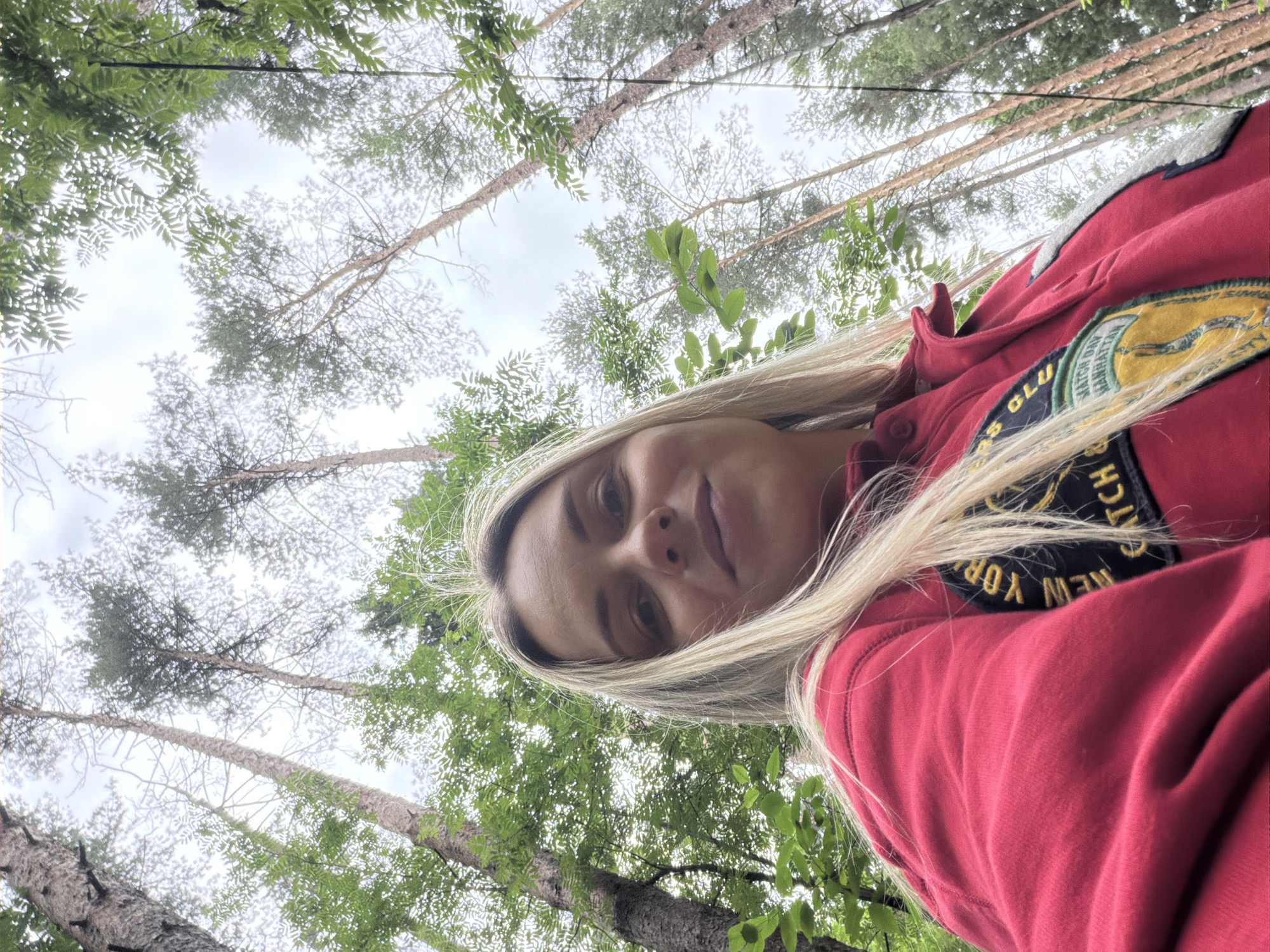 A low-angle, medium shot captures a young woman with blonde hair from the chest up, taking a selfie in a forest. She is wearing a red sweatshirt with patches on the chest. Her face is turned slightly towards the camera, with her head tilted back. The background consists of tall, slender trees reaching towards an overcast sky. The foliage of the trees is dense and green, with some bare branches visible. The image appears to have been taken in the city of Votkinsk, Russia.