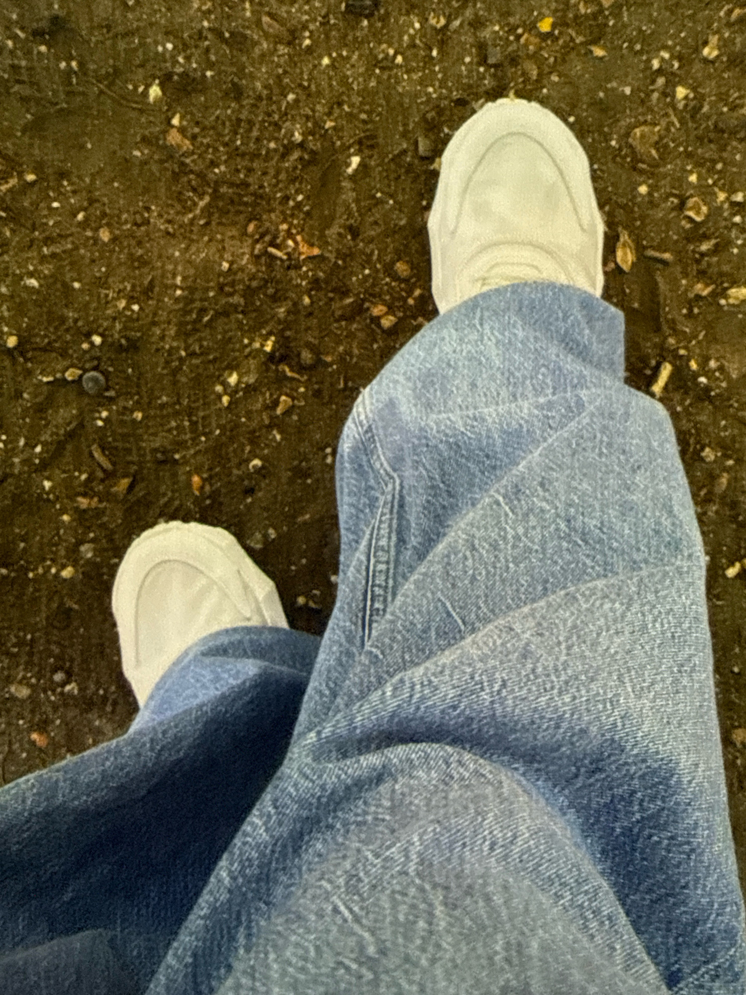 The image shows a top-down view of a person's legs and feet. The person is wearing light-colored sneakers and blue denim jeans. The jeans are creased and folded, indicating the person is likely sitting or resting. The ground beneath is dark and appears to be a mix of dirt, small stones, and possibly some dried leaves or debris. The overall impression is casual and outdoors. The context of Basildon, United Kingdom, is not directly discernible from the visual information in the image alone.