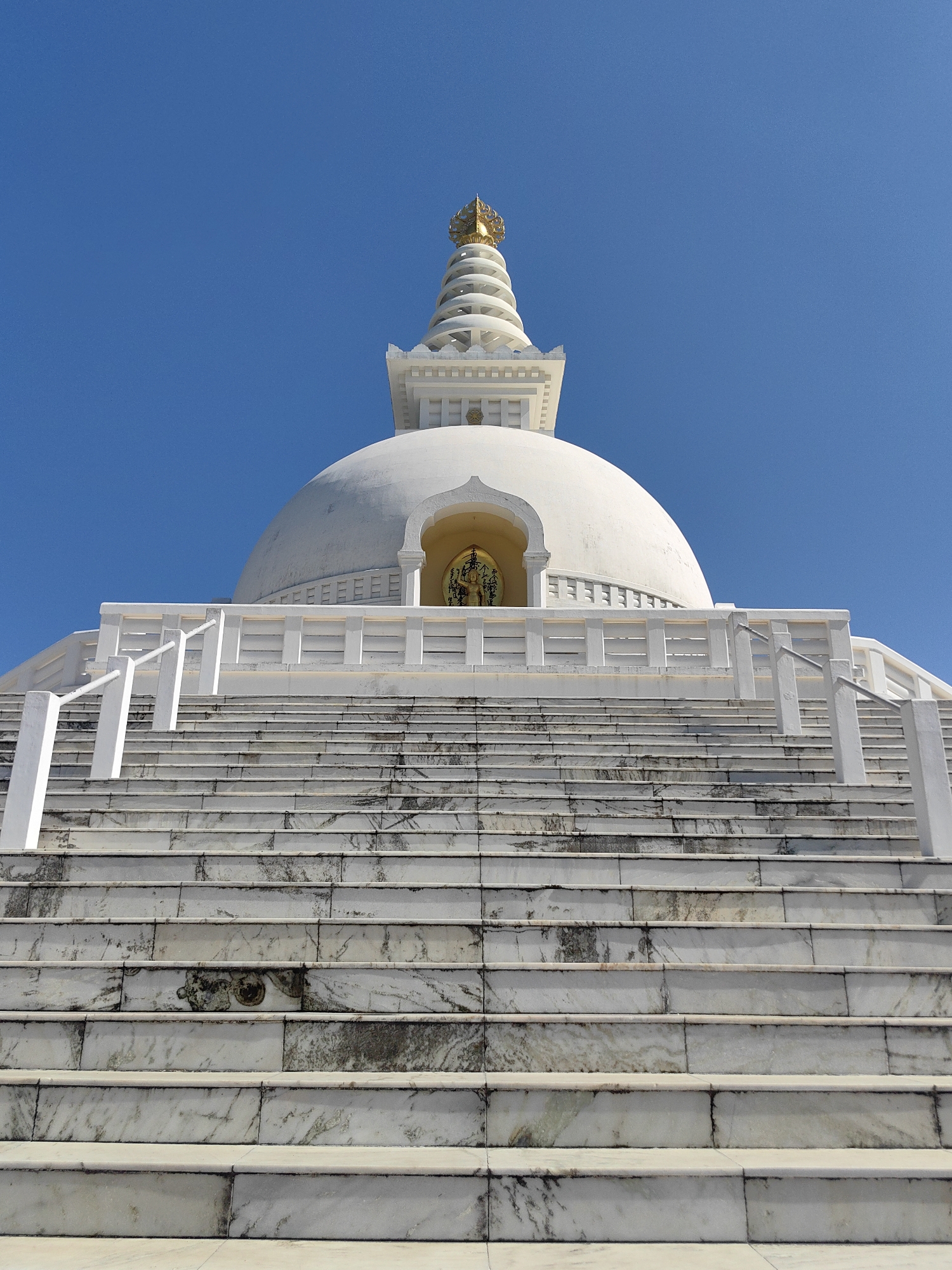 This is a low-angle, eye-level, medium shot of the World Peace Pagoda in Lumbini Bazaar, Nepal, under a clear blue sky. The white stupa, with a golden spire at its apex, dominates the upper portion of the frame. A grand marble staircase with white railings leads up to the base of the structure. A golden statue of Buddha is visible within an arched alcove at the base of the stupa. The marble steps show some signs of age and wear, with darker veins and smudges on their surfaces. The scene is tranquil and majestic, with no people or animals present.