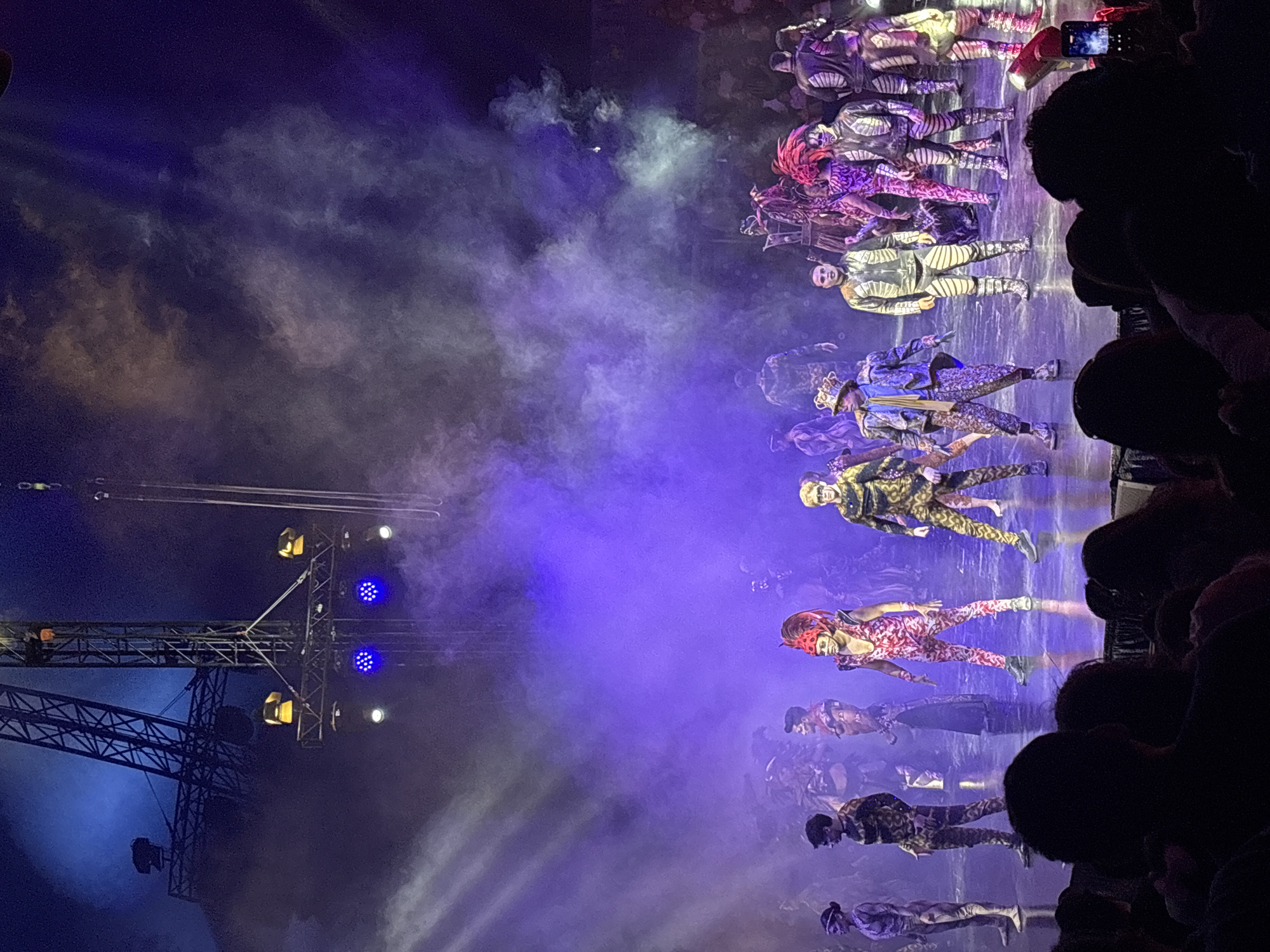 The image depicts a performance taking place on a stage, possibly a circus or theatrical event, in San Miguel, Peru. The stage is illuminated by dramatic purple and white lighting, with significant amounts of atmospheric fog or smoke present, particularly in the upper half of the image. Several performers, dressed in elaborate and colorful costumes, are spread across the stage. Their outfits appear to be themed, with some wearing patterned, form-fitting suits and others adorned with feathered headdresses and ornate headwear. One performer in the center foreground wears a bright red and white patterned outfit and a prominent red headdress. Another performer to the right of center is in a gold and black striped suit. In the background, a complex industrial-looking structure, possibly a crane or stage rigging, is visible against a dark, possibly night sky. The audience is partially visible in the foreground, appearing as dark silhouettes. One audience member in the lower right corner is holding up a smartphone, likely recording or photographing the performance. The overall scene conveys a sense of spectacle and theatricality.