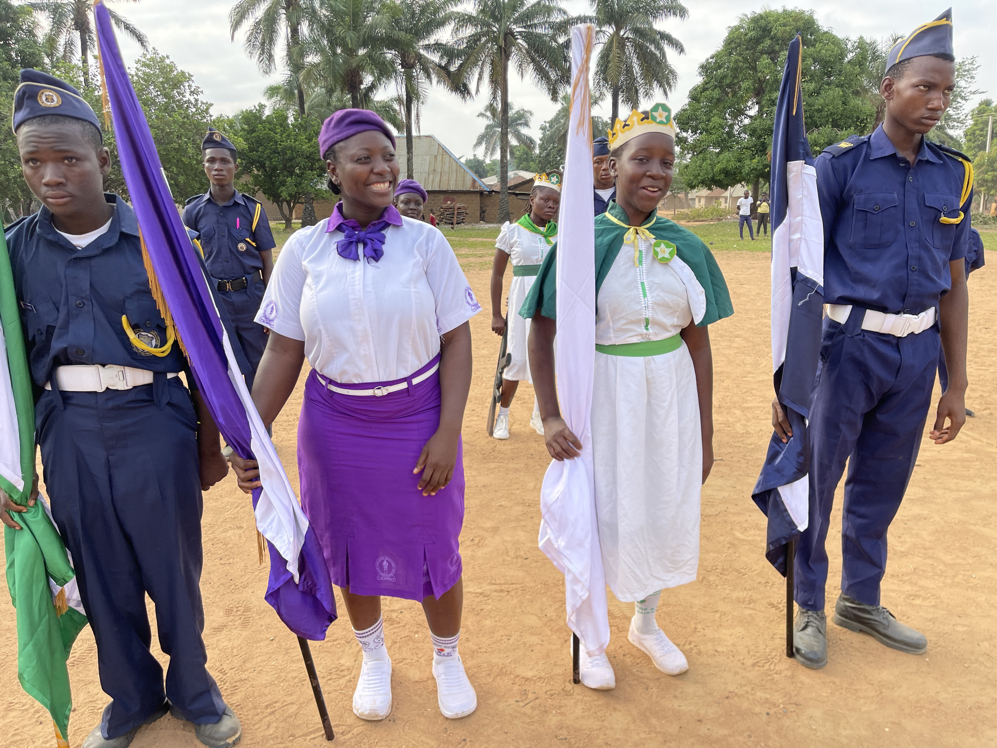 In a seemingly rural area of Tabu, Nigeria, a group of people, mostly young, are standing on a dirt-covered field. The individuals are clad in uniforms, suggesting they belong to an organization or group.
Two young men on the left and right of the frame are in matching blue uniforms with white belts and caps, holding flags with green, white, and purple colors.
Two young women stand in the foreground, one wearing a white blouse, purple skirt, and matching beret, and the other in a white dress with green accents and a crown. Both carry flags, one predominantly white and the other purple.
Several other uniformed individuals are visible in the background. The background also features a variety of trees, including palm trees, and rudimentary buildings. The sky overhead is overcast, providing diffused lighting.