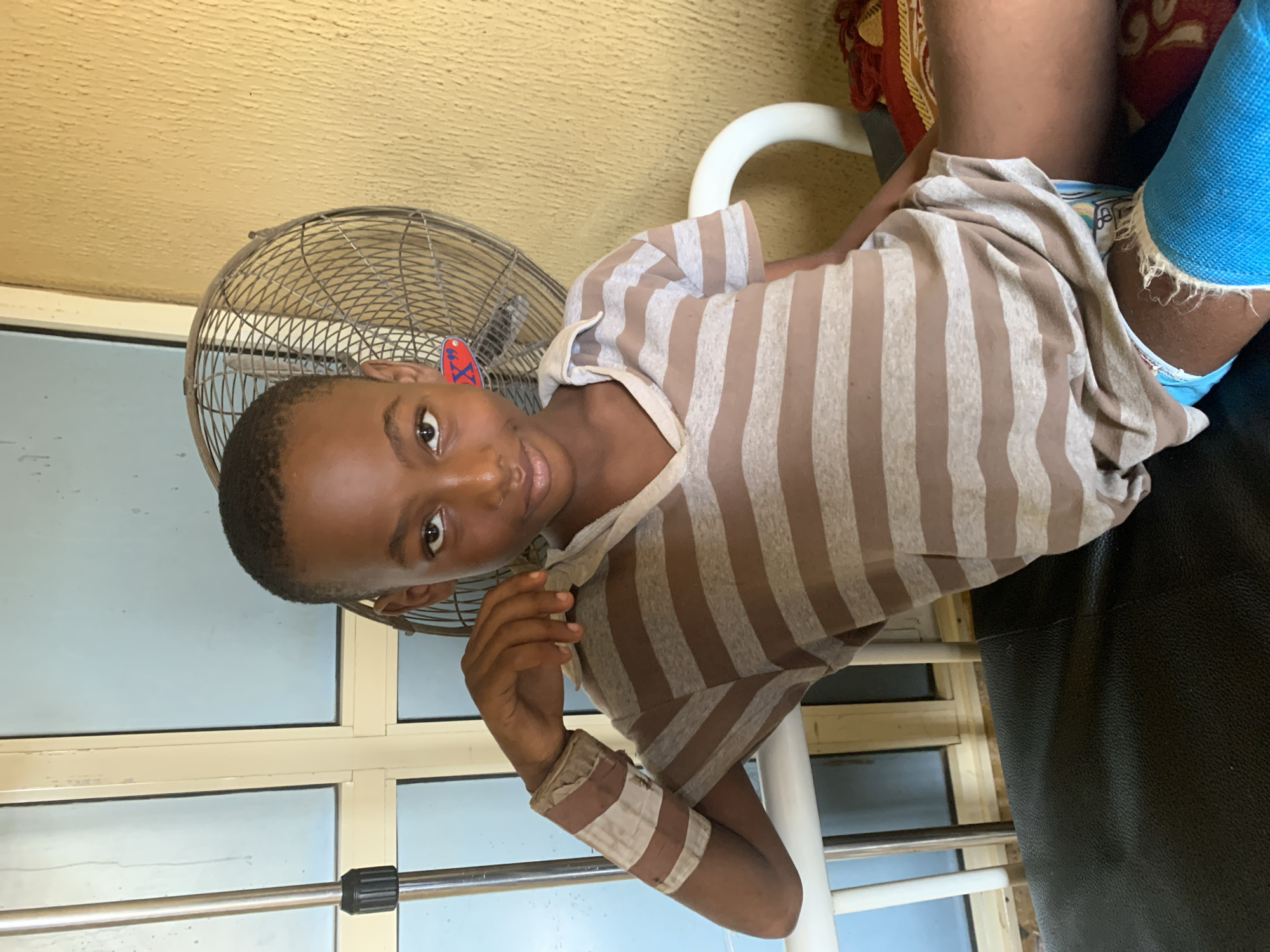 This media file captures a close-up, rotated shot of a young boy sitting down. He is wearing a light brown and white striped t-shirt, with a bandage on his right forearm. His left leg is encased in a bright blue cast, with the edges appearing frayed. The boy's expression is neutral, and he is looking directly at the camera.
Behind him, a metal fan with a circular grill is visible, and beyond that, a wall with a textured, yellowish surface. A portion of a white chair or support structure is also in the frame. The background also shows windows with pale blue panes, suggesting an indoor setting.
The overall scene appears to be a casual, possibly domestic environment. Given the location context of Shagamu, Nigeria, and the presence of a cast, it's plausible this image was taken during a recovery period. No specific time of day or weather conditions can be inferred from the image. There is a red sticker with what appears to be text on the fan, but it is too blurry to read.