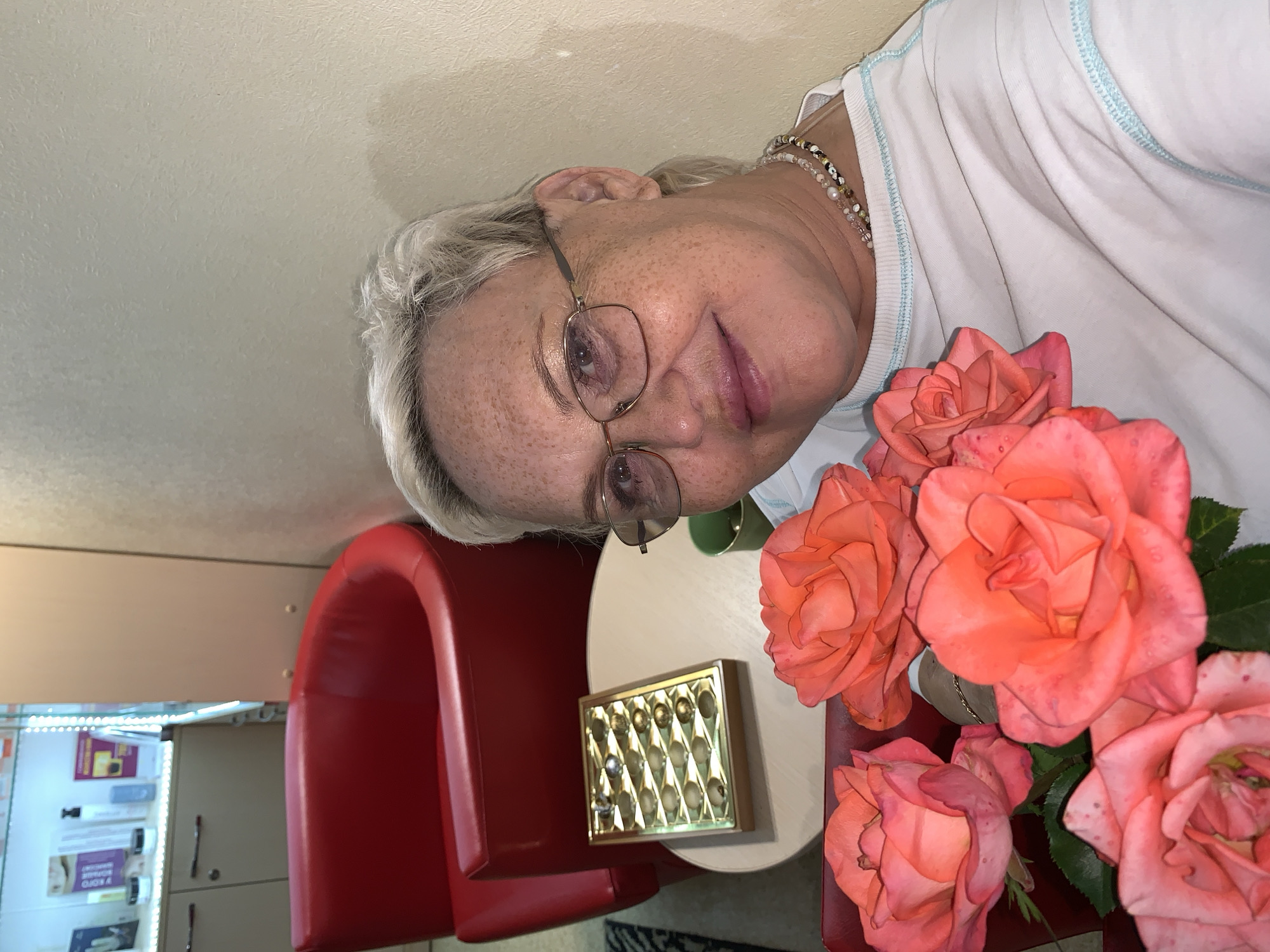 A woman with grey hair and glasses is holding a bouquet of coral-colored roses. She is wearing a white t-shirt with light blue trim and a beaded necklace. The woman is seated in front of a red chair and a round table. On the table, next to the roses, is a golden rectangular object with a diamond pattern, possibly a jewelry box. In the background, there are shelves filled with products, suggesting a retail or salon setting. The lighting is bright, indicating it is daytime. The woman's expression is neutral.