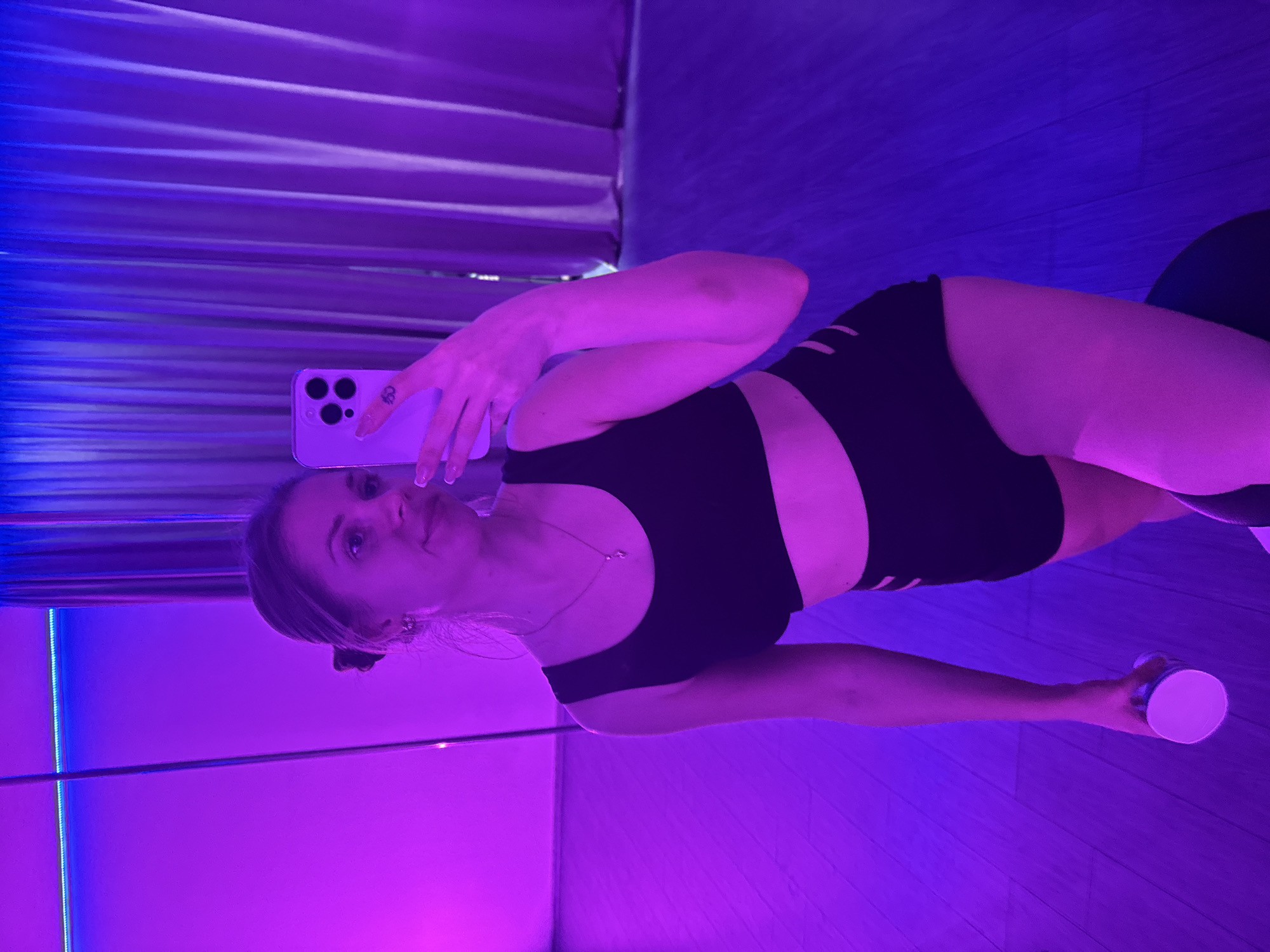A woman takes a selfie in a room with a pole. She is wearing a black sports bra and black shorts. The room has a blue and purple lighting, and the floor appears to be made of wood. The location may be in the city of Batumi, Georgia.