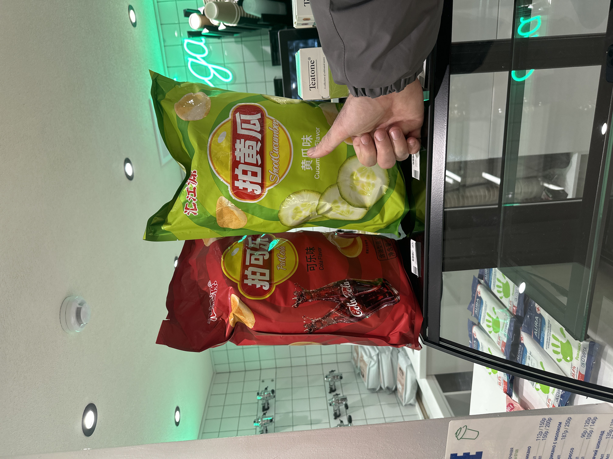 Here's a forensic description of the image:
The image depicts a close-up view inside a store, possibly a convenience store or market. Two bags of Lay's potato chips are prominently displayed behind a glass counter or display case. The bag on top is green and labeled "Shoot Cucumber" with cucumber slices depicted on the packaging, indicating a cucumber flavor. The bag underneath is red and labeled "PatCola", with images of Coca-Cola bottles, signifying a cola flavor. A hand, presumably belonging to a customer, is pointing at the cucumber flavored chips.
Behind the chips and above the counter, a neon green sign is partially visible. A box from "Teatone" is also visible on the counter.
Below the potato chips, inside the display case, are packages of Aura antibacterial wipes. Some price labels are visible at the bottom of the image, written in Russian.
The background includes a white tiled wall with embedded lights.
The location is Unknown, Unknown.