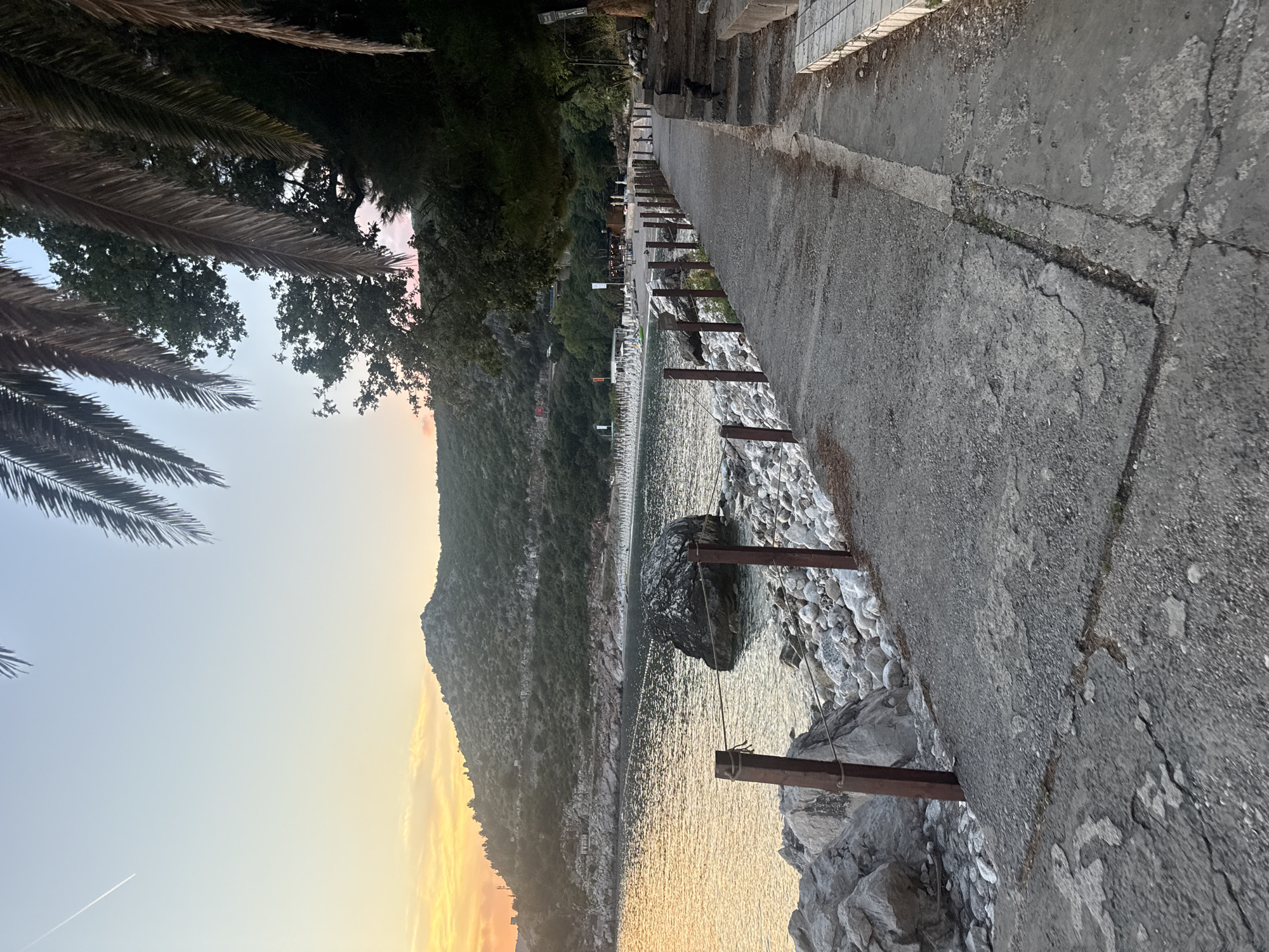 The image captures a view of a paved walkway adjacent to a rocky shoreline in Kamenovo, Montenegro, during a sunset. The walkway, made of concrete, shows signs of wear and tear, with cracks and uneven sections. A wooden fence with metal posts and ropes lines the edge of the walkway, separating it from the rocky beach. The sea is calm, reflecting the warm hues of the setting sun, with a large boulder visible in the water near the shore. Beyond the beach, a line of white beach chairs and umbrellas suggests a developed tourist area. The background features a lush, green hillside with winding roads. Palm trees frame the upper right portion of the image, their fronds casting shadows. The sky is a gradient of pale blue and soft orange, indicating the end of the day. No people are prominently featured in the scene.