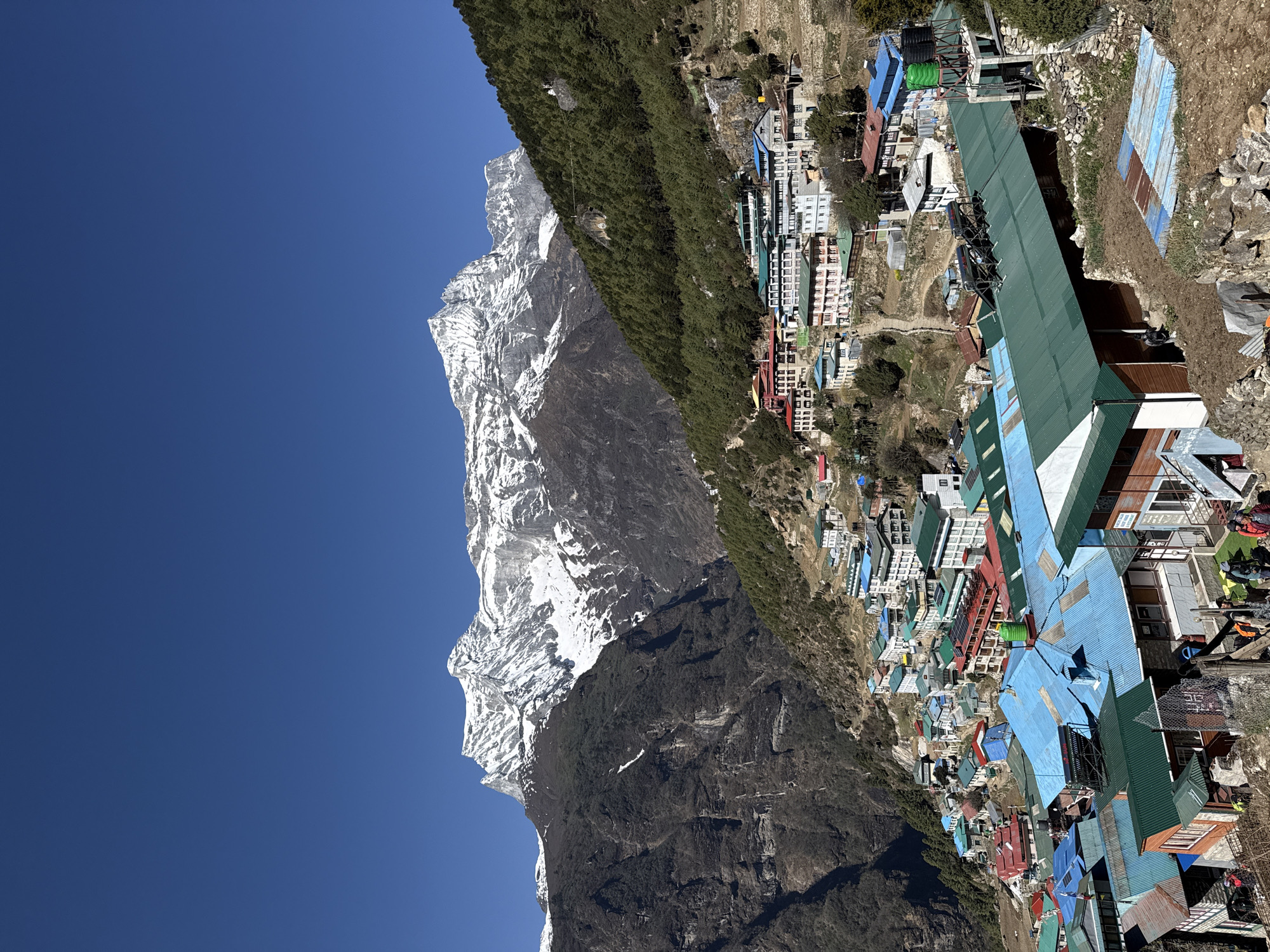 The image captures a mountainous vista near Namche, Nepal. A steep, snow-capped mountain looms on the left, its rocky face transitioning into lush, green vegetation at lower altitudes. To the right, a densely packed settlement unfolds, with buildings exhibiting varied colors, primarily blue, green, and red roofs. The structures appear to be closely clustered, suggesting a compact village layout. The sky overhead is a clear, vibrant blue, offering a stark contrast to the rough terrain below. The scene is bathed in sunlight. The image showcases the unique juxtaposition of natural landscapes and human settlements in the Himalayan region.
