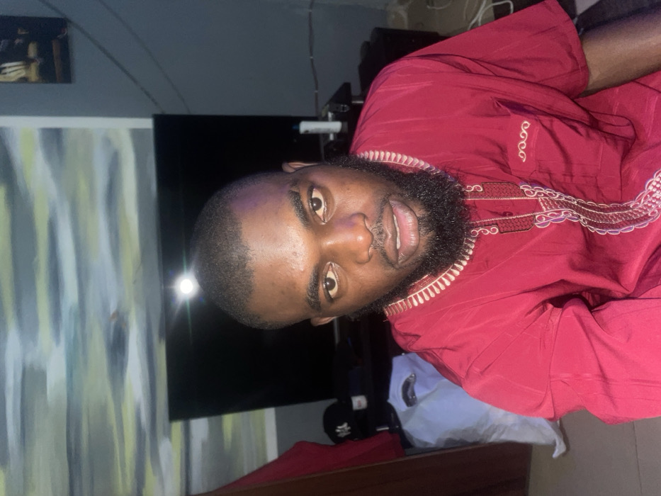 A dark-skinned man with a beard and short hair is the focus. He wears a red garment with decorative trim. A painting with vertical lines is on the wall behind him. To the right is a black television screen. Wires are visible near the top of the frame. There are other items in the background but are too indistinct to identify. The overall lighting is soft.