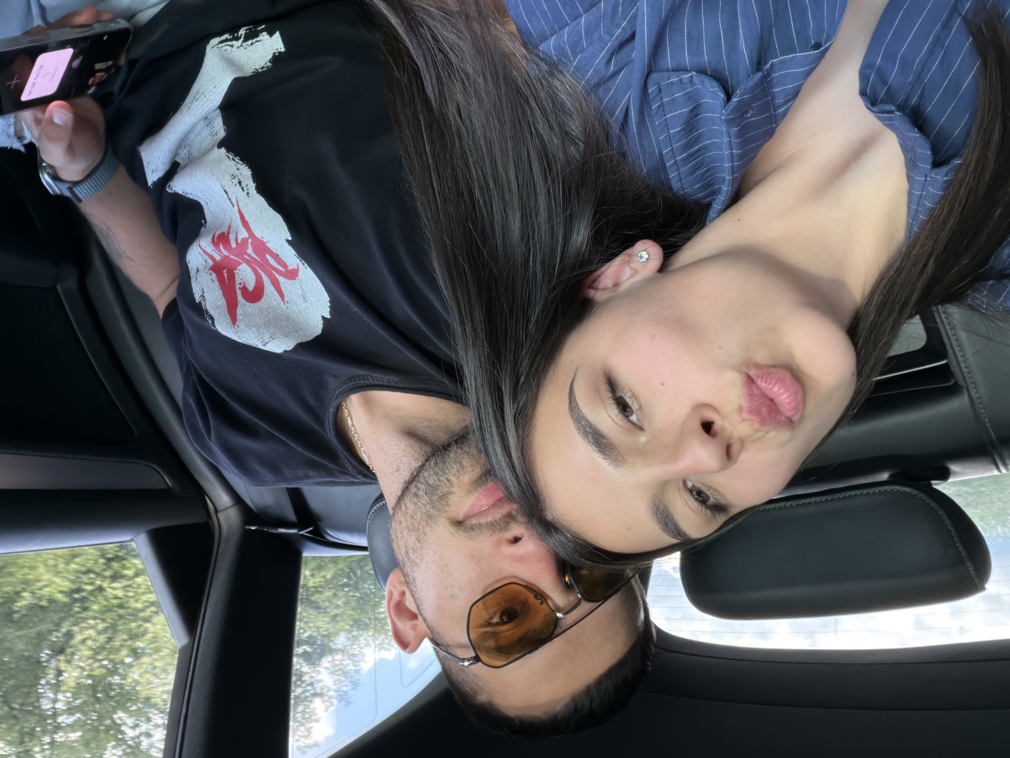 Two individuals are in the interior of a vehicle. One individual wears sunglasses and a gold chain. The other individual is wearing a blue pinstripe shirt. A cell phone is visible in the image. Outside, trees and sky can be seen. The location is unspecified.