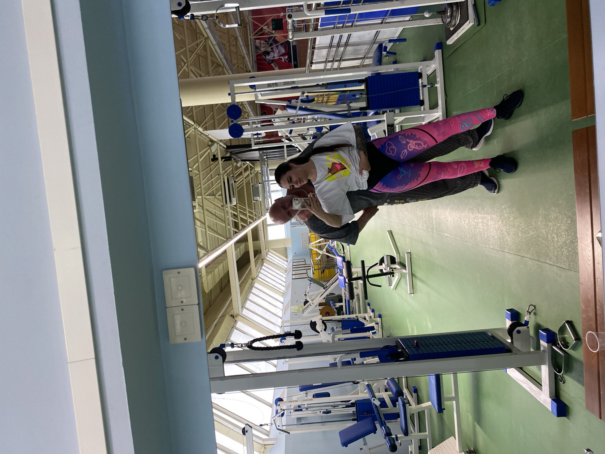 A snapshot in a gymnasium in Chaikovsky, Russia, captures a man and a woman taking a selfie, with gym equipment scattered throughout the scene. The woman, positioned in front, dons a white t-shirt and pink and purple patterned leggings, her long hair contrasting with the bald head of the man standing behind her. He is wearing a gray t-shirt. The gymnasium's green floor is surrounded by a variety of blue and white weight machines. The room is lit by natural light coming through the ceiling windows. A large banner hangs in the background.
