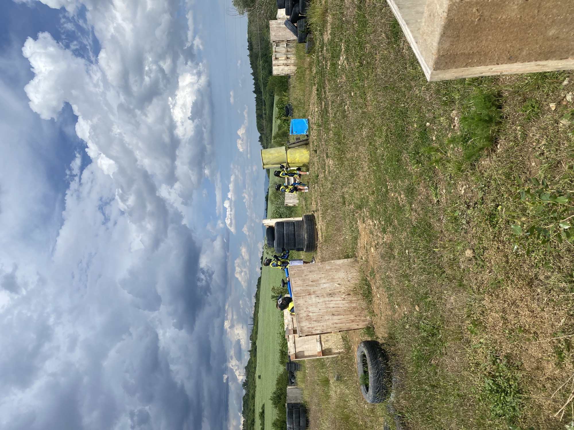 This image captures an outdoor paintball or airsoft field, likely located near Sofia, Bulgaria, under a partially cloudy daytime sky. The scene is dominated by a vast expanse of ground, covered in dry grass with interspersed greener patches, and a dynamic sky filled with large, white and grey cumulus clouds against a backdrop of blue.
Several individuals, presumably players, are visible in the mid-ground, engaged in the game. They are wearing protective gear, including helmets and bright yellow-green vests or jackets over darker clothing, indicating a focus on safety during the activity. The players are strategically positioned behind various forms of cover, which are scattered across the field. These barriers include structures made from wooden pallets or crates, tall stacks of black vehicle tires, and a distinctive yellow barrel-like object. A bright blue tarp or large object also stands out as a potential cover element.
In the background, beyond the immediate playing area, stretches a landscape of lush green fields, possibly agricultural, and a dense line of trees forming a horizon, suggesting a rural or semi-rural setting. The lighting indicates a bright, sunny day, despite the presence of significant cloud cover. No discernible text is visible within the image.