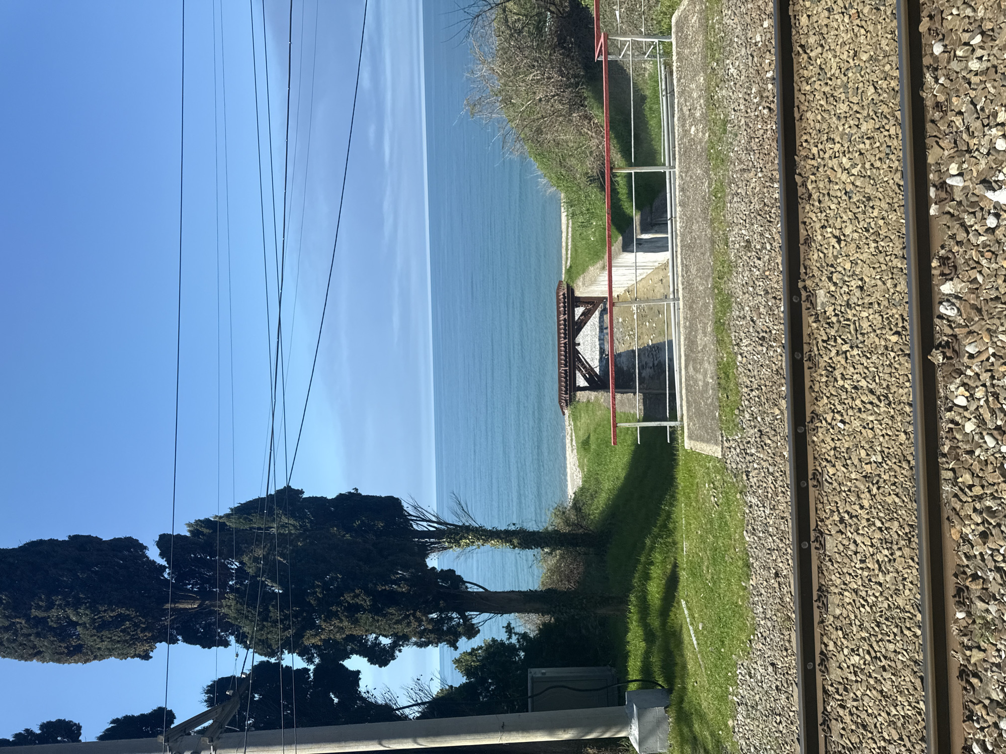 The image shows a view from beside a railway track, looking out towards a body of water. The railway track is visible on the right side of the image, running parallel to the frame. To the left of the tracks, there is a grassy area. Beyond this area, there is a drop down to the water's edge, where a small shelter structure with a red roof and metal railings can be seen. The water extends into the distance, taking up a large portion of the image. The sky above is clear and blue. On the left side of the image, there are trees and power lines running vertically. The location appears to be coastal, with a railway track running alongside the water.