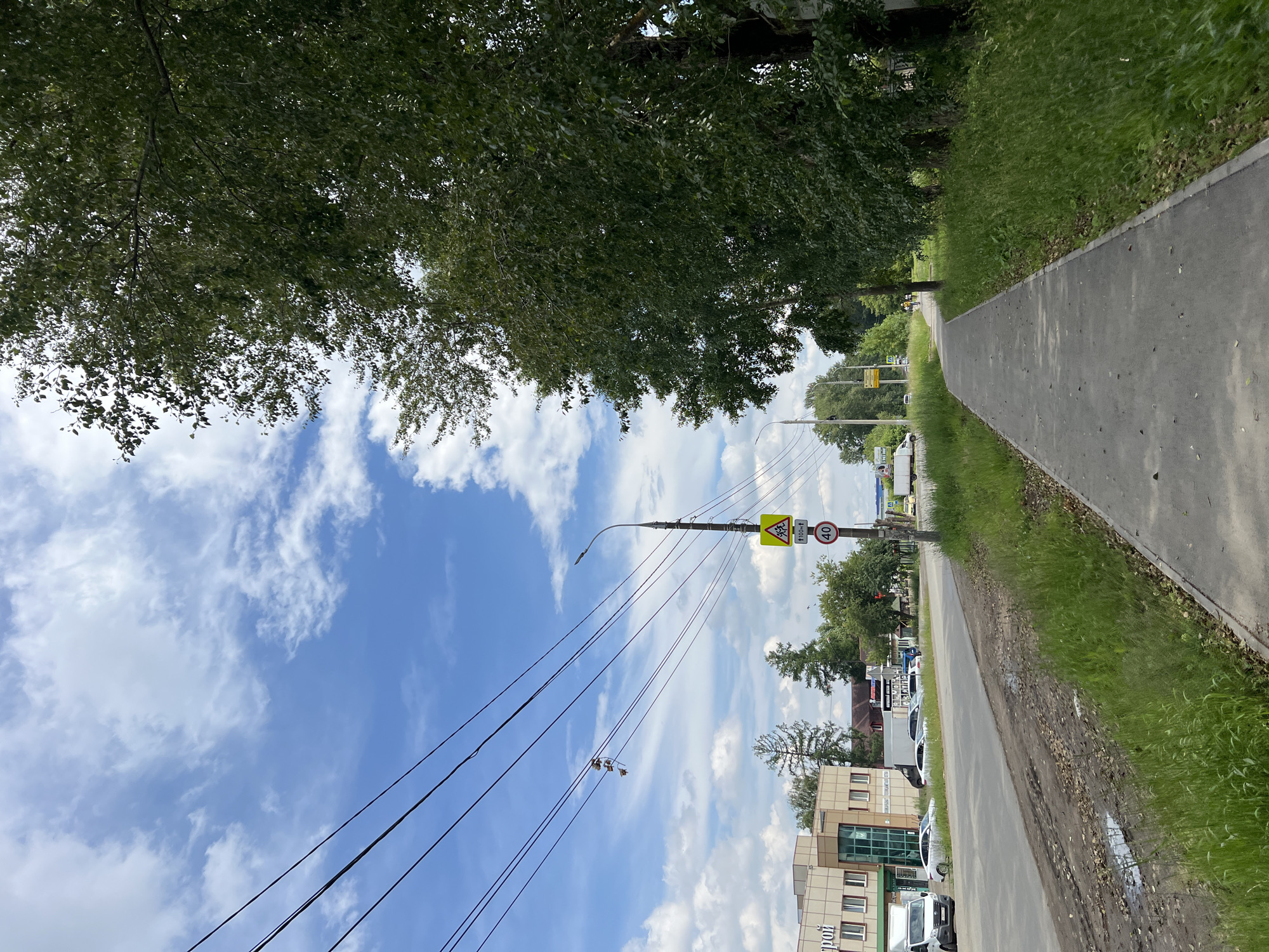 This image depicts a street scene in Izhevsk, Russia. A paved sidewalk runs along the right side of the frame, bordered by tall, leafy green trees and overgrown grass. To the left of the sidewalk is a road with several cars parked and driving. On the sidewalk, a pedestrian is visible in the distance, walking away from the camera. Overhead, power lines stretch across the sky, and a streetlight stands on a pole with traffic signs indicating a pedestrian crossing and a speed limit of 40 km/h. A modern building with windows and signs can be seen on the left side of the road. The sky is blue with scattered white clouds.
