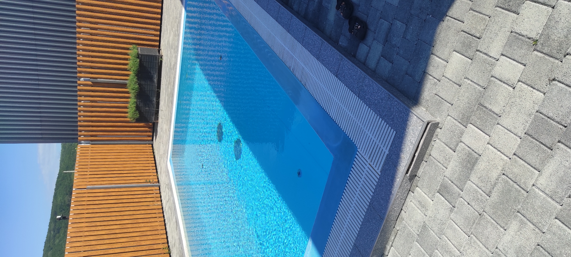 This image depicts a clean, modern outdoor swimming pool area on a sunny day, likely in or near Tulskiy, Russia. The pool itself is a bright, clear blue, reflecting the sky. It is surrounded by a paved patio made of grey interlocking bricks. A low, slatted drainage system runs along the edge of the pool. To the left and behind the pool, a light brown wooden fence with vertical slats encloses the area. Beyond the fence, a lush green hillside is visible under a bright blue sky. To the right of the pool, a dark grey, corrugated metal building stands. In the background, a rectangular black planter filled with green plants is placed against the fence, near the building. A pair of dark flip-flops rests on the paved area near the bottom right corner of the image. The scene is tidy and appears to be a well-maintained private residence or resort.