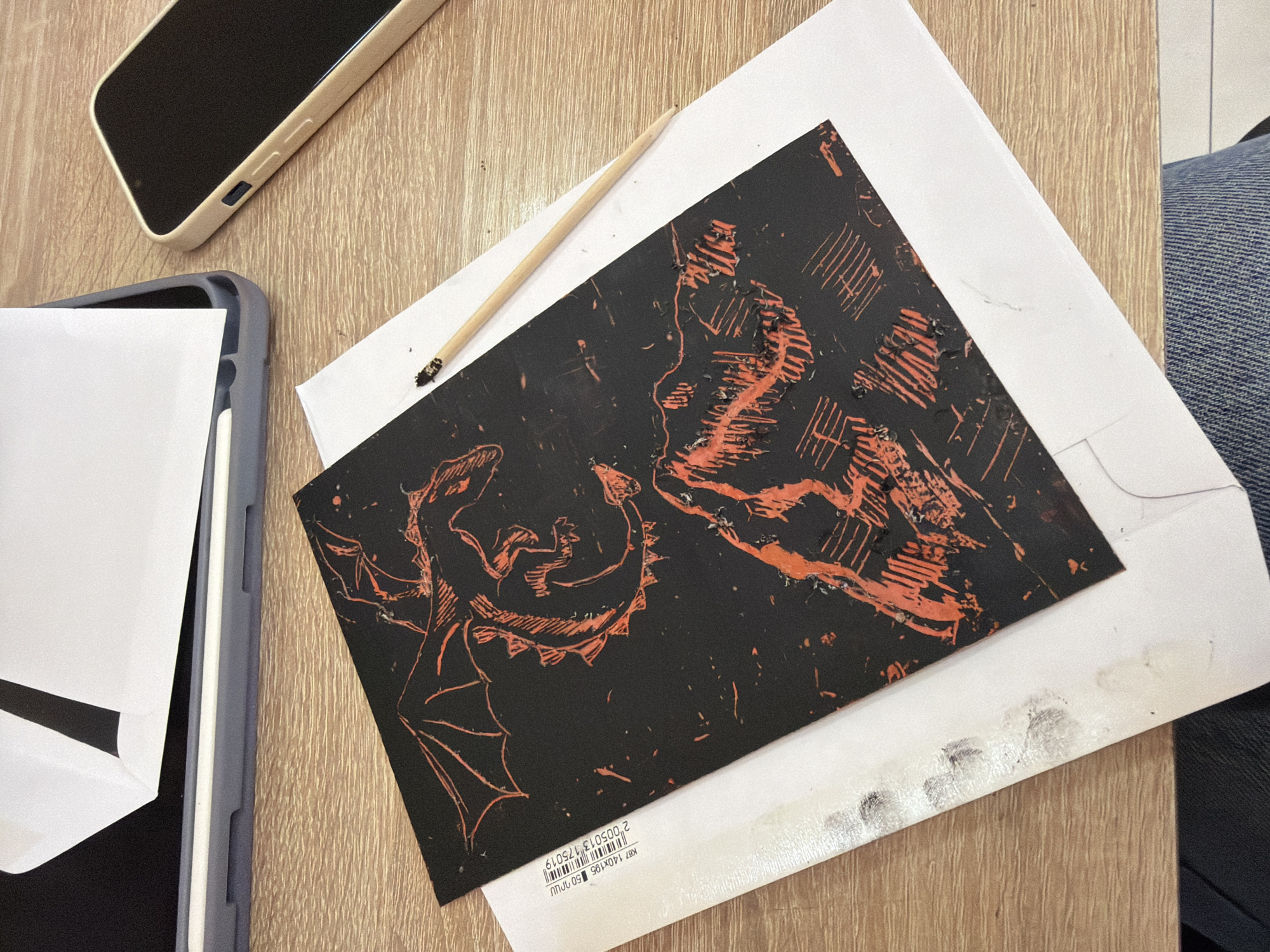 A close-up overhead shot depicts a workspace on a light-colored wooden surface. Visible items include a black-cased mobile phone, a tablet in a grey case partially visible with a white sheet of paper, and a linocut print depicting two dragons facing each other. The print is black with orange-red lines forming the dragon imagery. A small wooden stick, likely used in the linocut process, rests partially on the print. The print sits on a white envelope, with black smudges near the envelope's corner. A person's leg clad in denim is partially visible in the upper right corner. The scene is indoors, and the specific location within Yerevan, Armenia, cannot be determined based on the image alone.