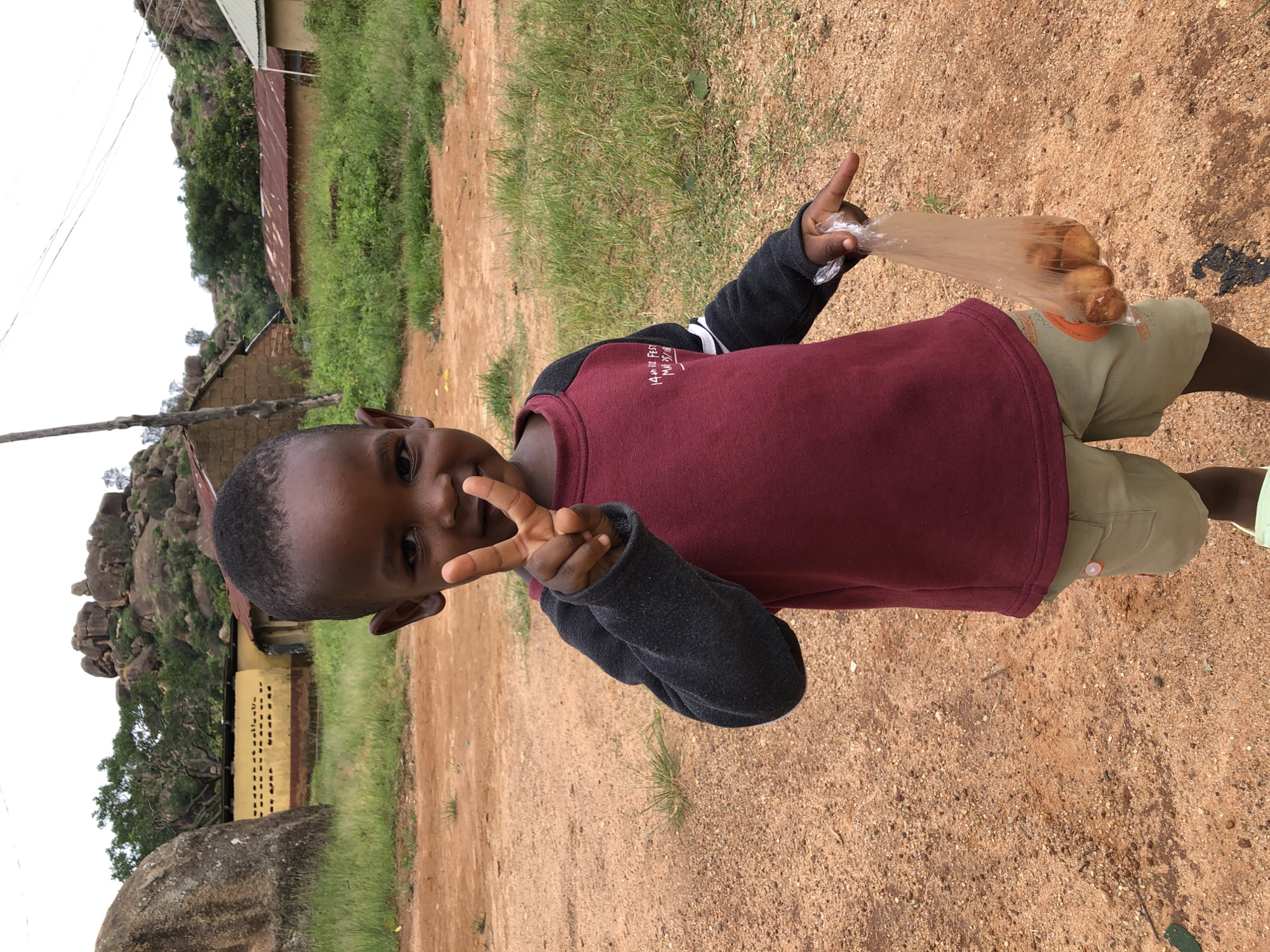 A young boy, appearing to be of African descent, stands outdoors in what seems to be a rural or semi-urban area. He is facing the camera and making a peace sign with his left hand. He is wearing a maroon and dark gray long-sleeved shirt and light brown shorts. In his right hand, he holds a clear plastic bag containing several round, fried food items, possibly puff-puff or similar snacks. The ground is covered in reddish-brown dirt and small stones, with patches of green grass visible. In the background, there are buildings with corrugated metal roofs and stone structures, suggesting a residential or village setting. The landscape is characterized by rocky hills and trees. The sky is overcast. This scene is consistent with the environment found in the city of Jos, Nigeria, known for its rocky terrain and blend of urban and rural settlements.