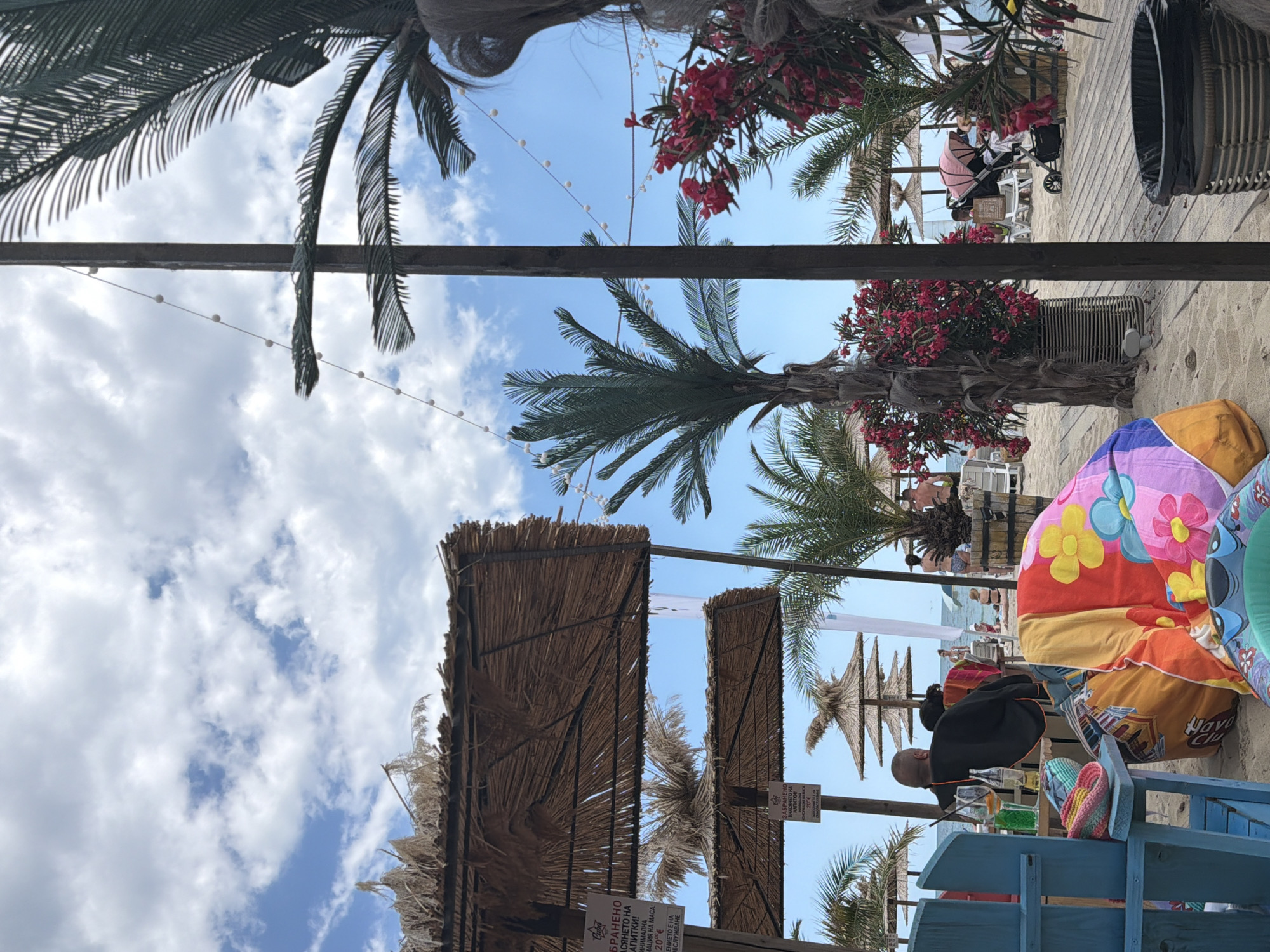 The image depicts a beach scene on a sunny day in Varna, Bulgaria, likely at a seaside resort. In the foreground, a person is seated in a blue chair, facing away from the camera, with drinks on a table beside them. Colorful beach blankets and bags are strewn on the sand. Several palm trees and straw-roofed umbrellas are visible, characteristic of beachside establishments. In the background, other beachgoers, sun loungers, and a baby stroller suggest a recreational area. The sky is partly cloudy, with a clear blue sky visible between the clouds. The overall setting is a relaxed and casual atmosphere typical of a beach vacation.