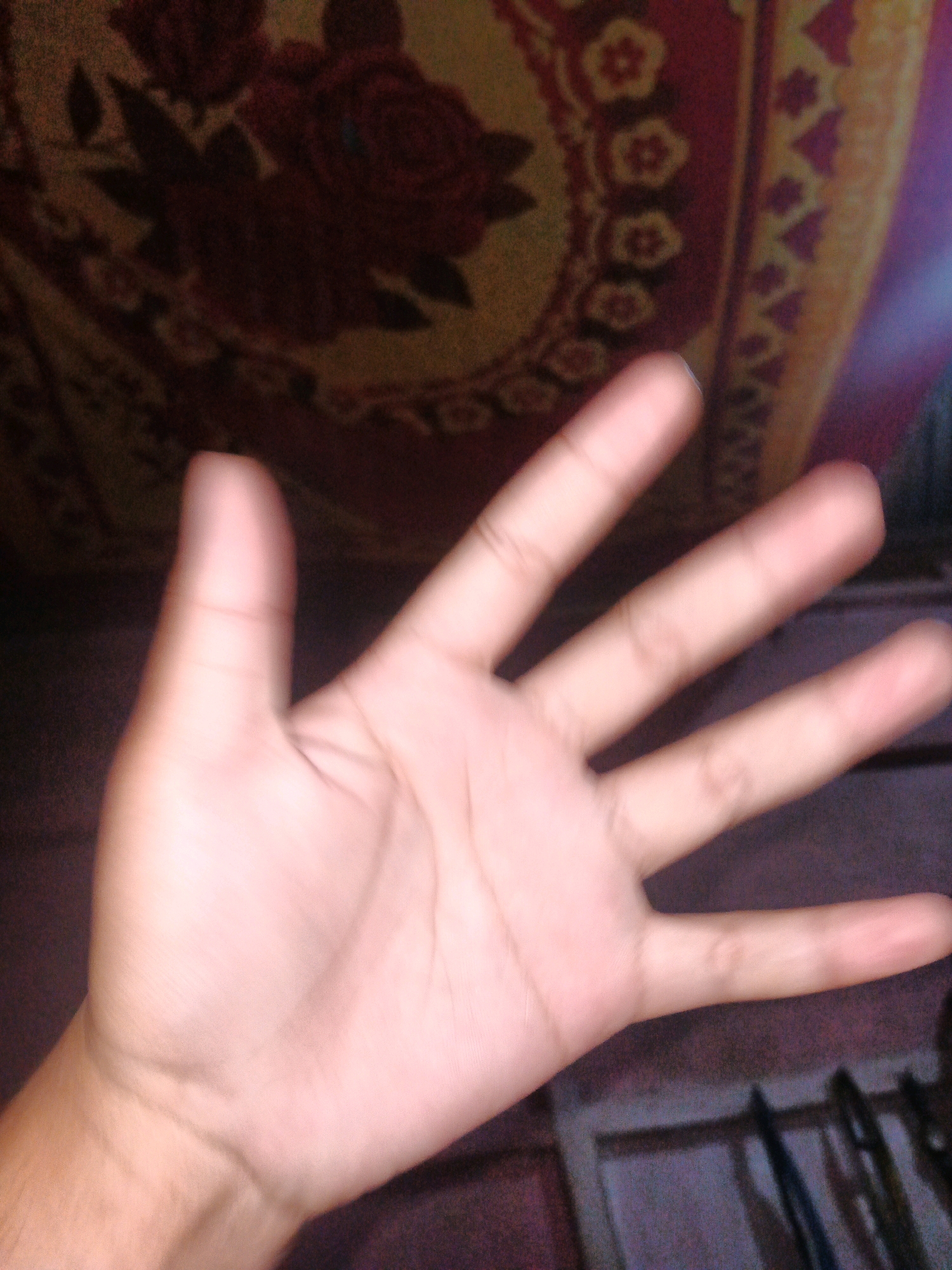 A person's left hand is open and facing the camera, with the palm and fingers clearly visible. The hand is positioned in front of a patterned cloth with red and yellow floral designs. The background is blurred, showing dark tones and a suggestion of vertical objects. There is no discernible location in Padengo, Indonesia.