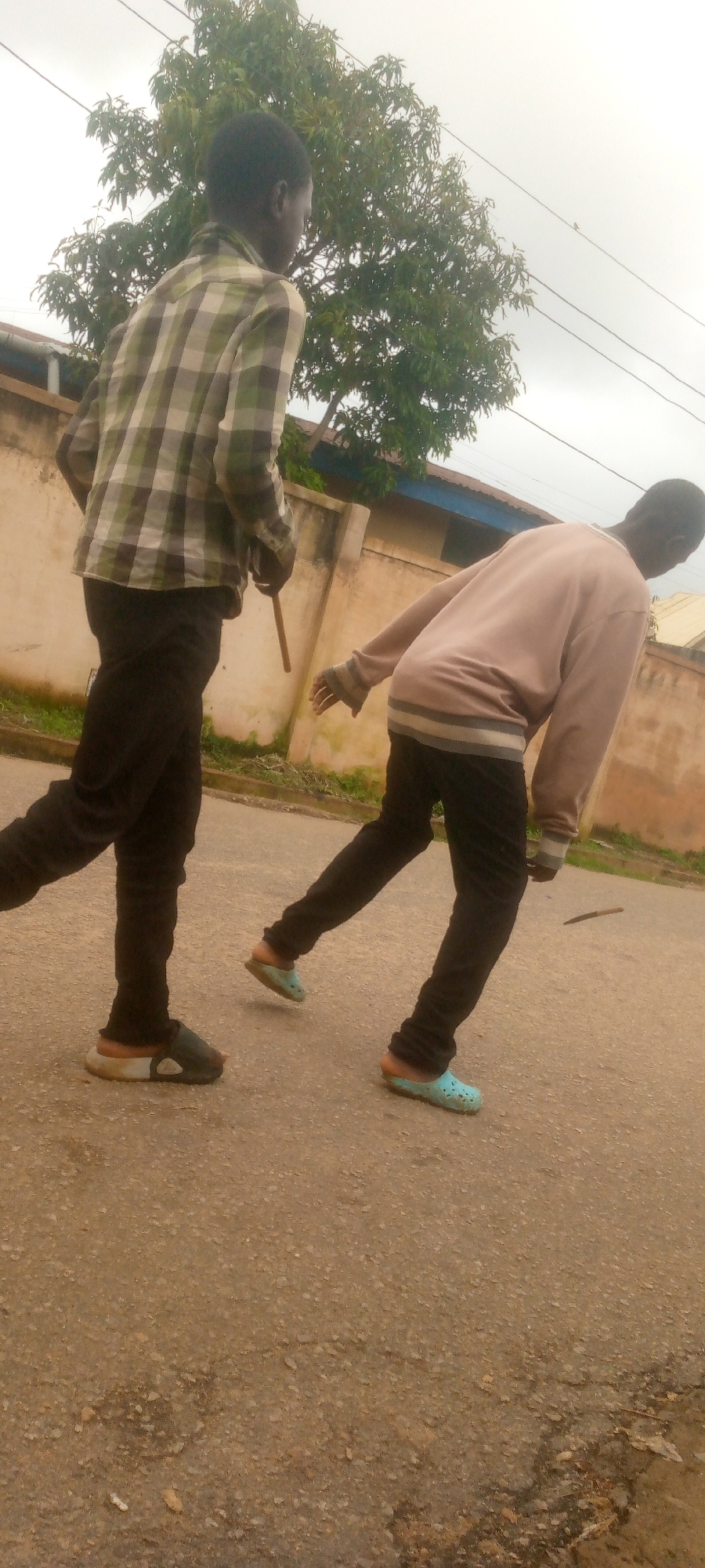 The image depicts two young males walking outdoors on a paved surface, possibly a road or sidewalk, in an urban environment. The male in the foreground on the left is wearing a green and white checkered long-sleeved shirt, black pants, and dark slip-on shoes. He appears to be holding a thin, dark object, possibly a stick or a cane, in his right hand. The second male, on the right, is wearing a light-colored long-sleeved shirt or sweatshirt, dark pants, and light blue slip-on shoes. He is bent slightly forward, with his right arm extended downwards. Behind the individuals, a low, light-colored wall runs horizontally across the frame, with a building partially visible beyond it. A large tree with green foliage is positioned behind the wall and the male on the left. The sky is overcast and pale. The overall scene suggests a casual outdoor activity or movement through a residential or less-trafficked area.