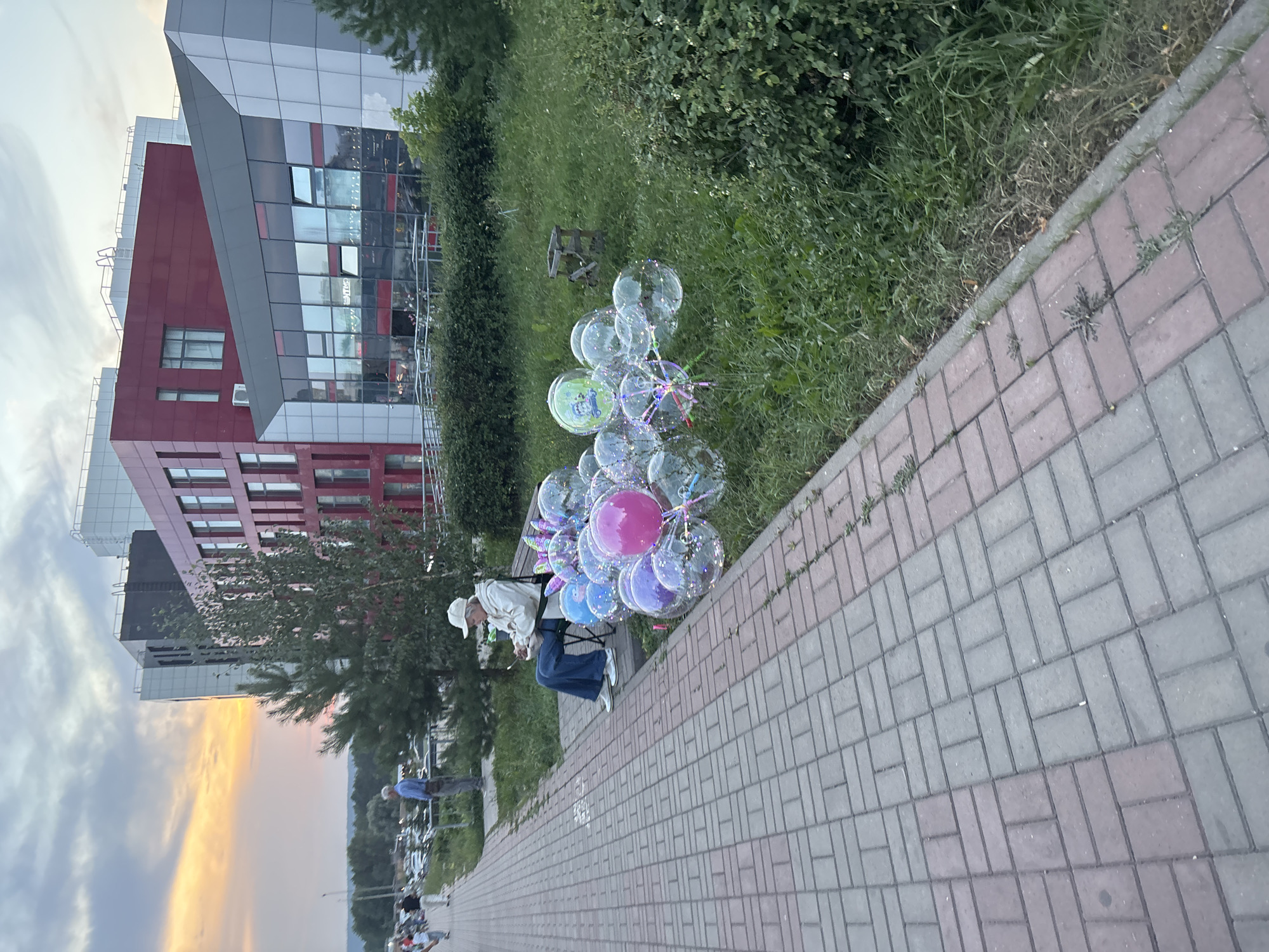 The image depicts a scene on a sidewalk in Naberezhnye Chelny, Russia, near a modern building with a red facade and large glass windows. A woman wearing a white hat, beige jacket, and blue jeans sits on a bench, appearing to be engaged with something in her hands. Beside her are a large cluster of clear bubble balloons, some with colorful glitter or confetti inside. The sidewalk is paved with red and gray bricks, and a strip of green grass borders it, leading to dense foliage and trees. In the background, other people are visible walking along the sidewalk and parked cars are present. The sky above is cloudy with a hint of orange from a sunset.